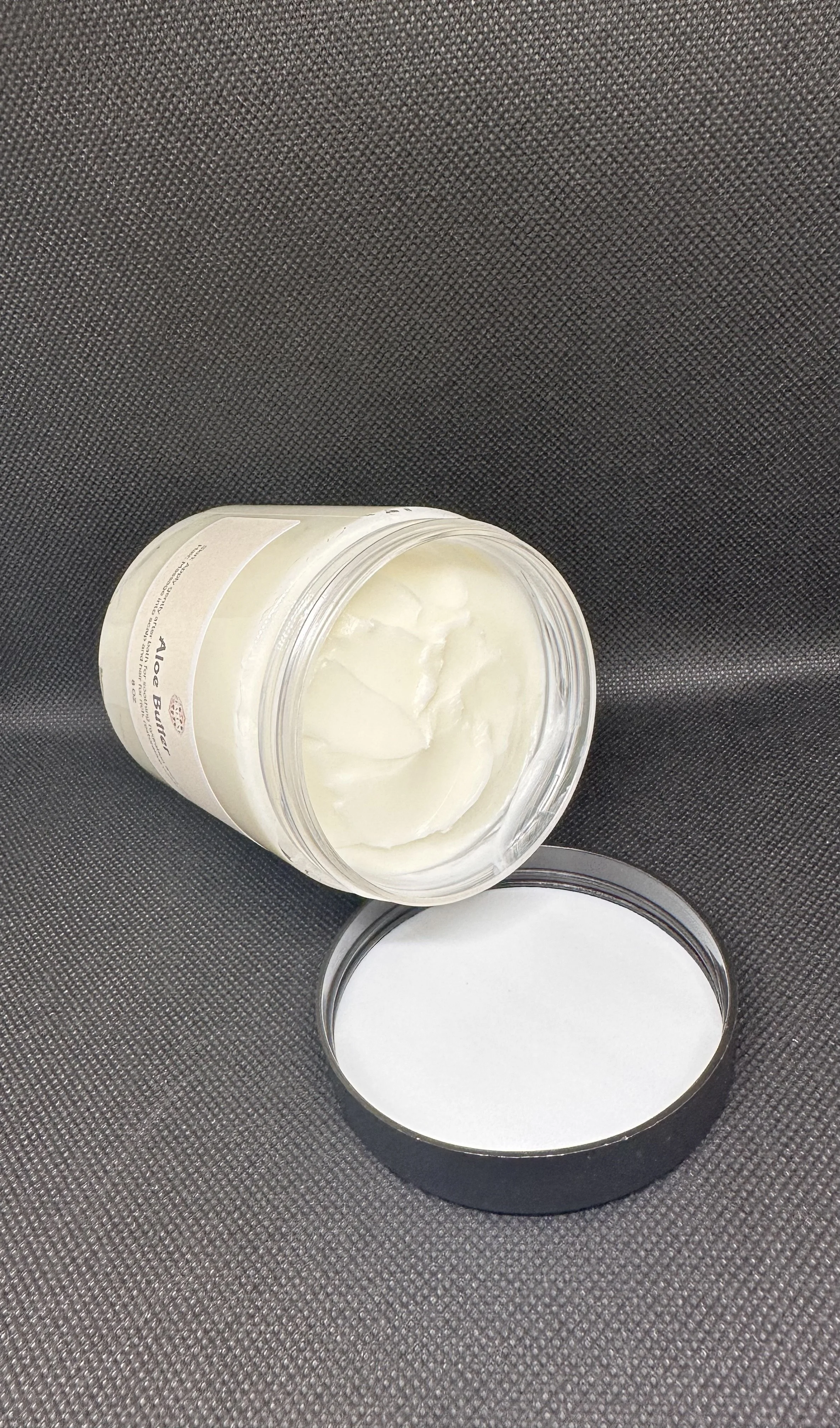 Open jar of white cream or lotion on dark textured surface with detached metal lid beside it.