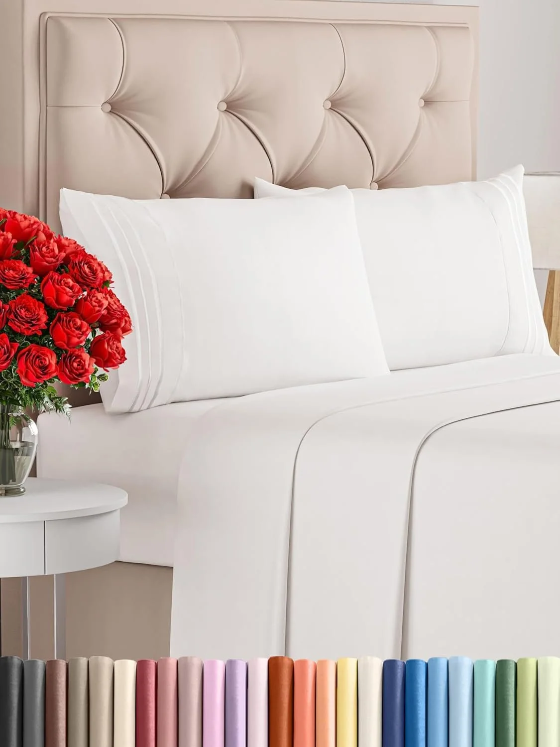 White bed with beige tufted headboard, white pillows, and a bouquet of red roses on a white side table, with colorful spines of books or fabric samples at the bottom of the image.