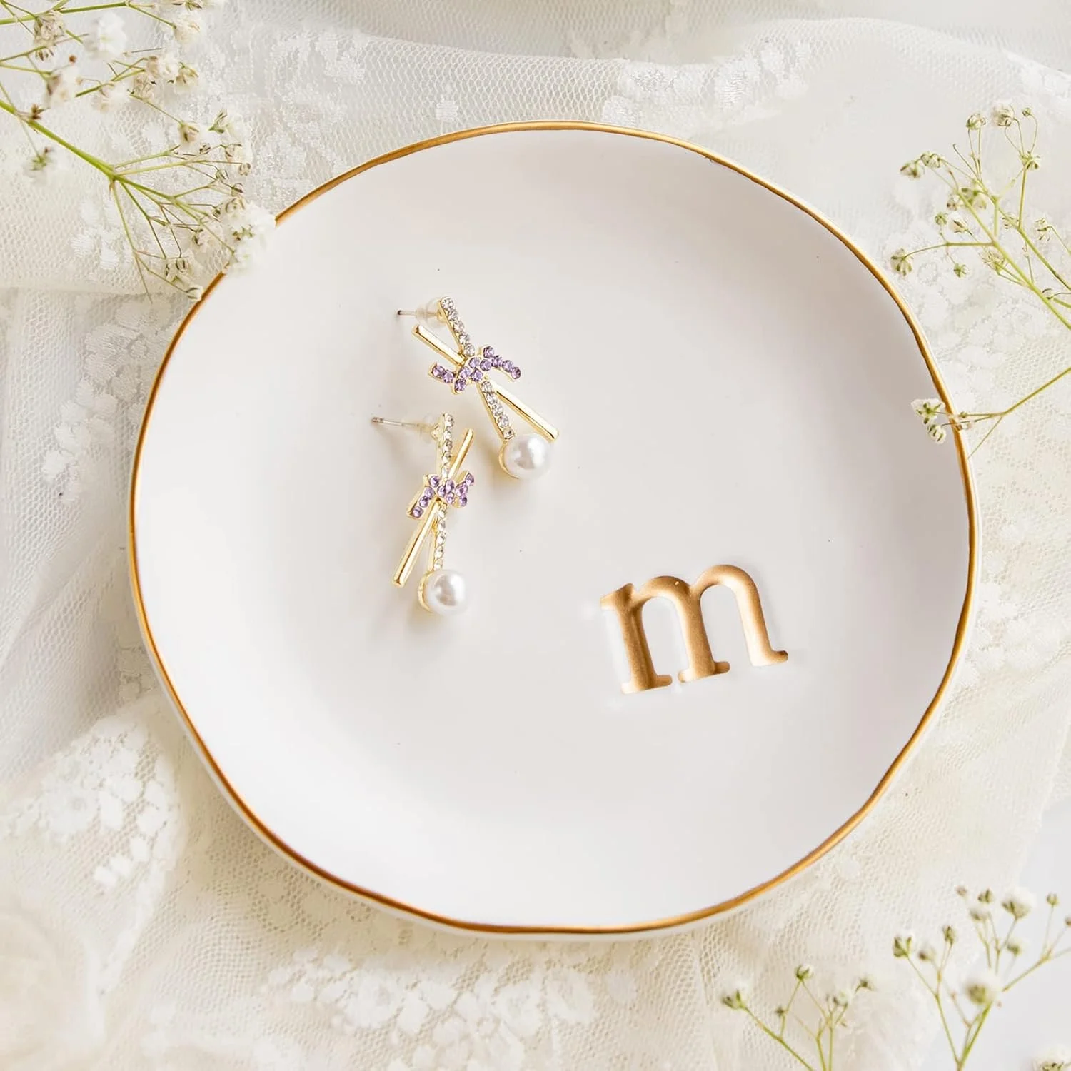 A white ceramic jewelry dish with a gold rim, holding a pair of gold earrings with purple and clear rhinestones, pearl drops, and purple bow accents. The dish has the letter 'm' cut out in gold. The dish is placed on a lace surface with small white flowers around it.