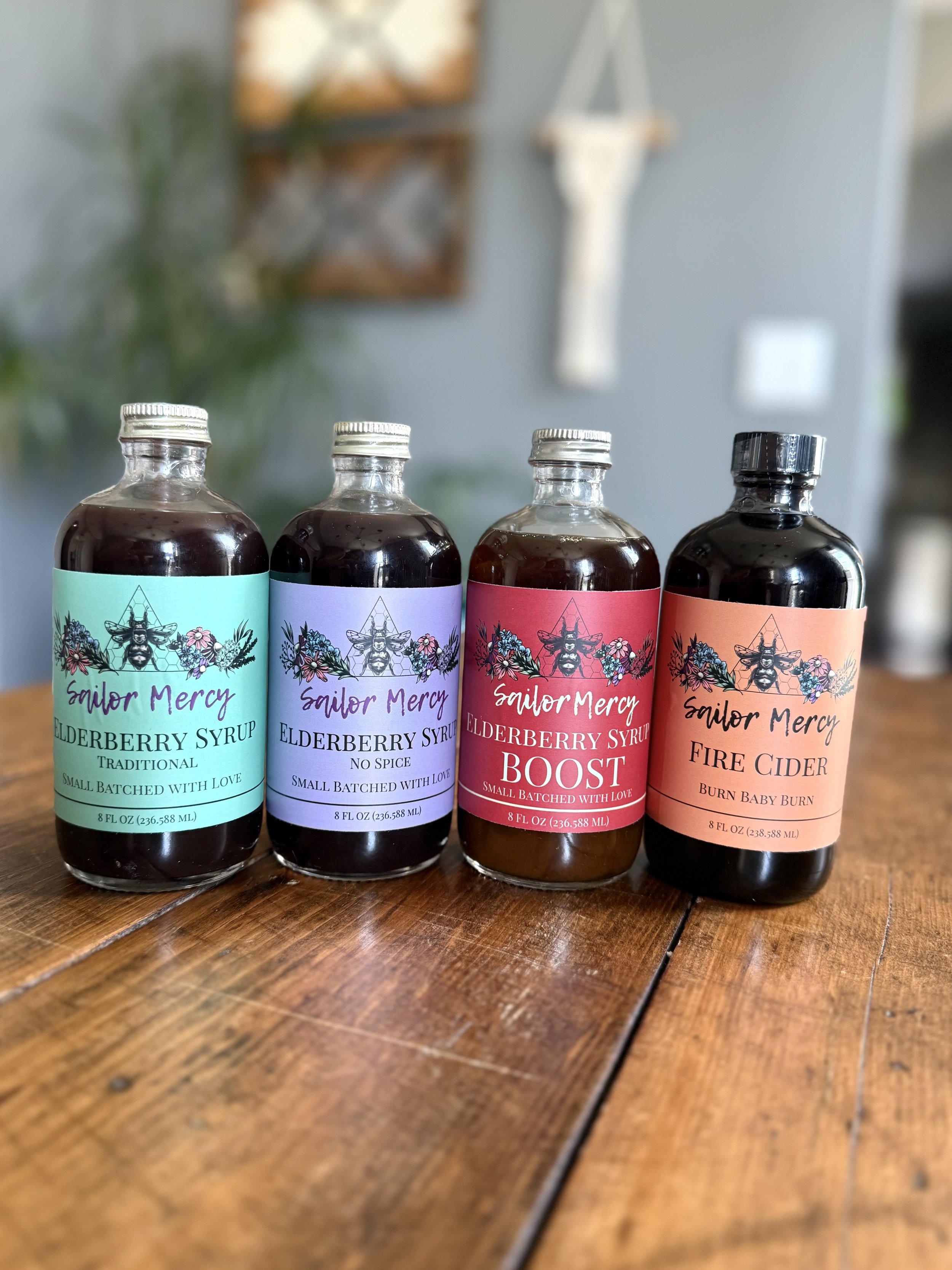 Four bottles of craft beverages with colorful labels on a wooden surface, backdrop of a cozy interior with wall decor and a plant.