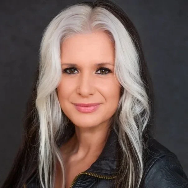 A photo of award winning director Lisa Ovies. She has long, wavy, black and white hair, wearing a black leather jacket, smiling at the camera against a dark background.