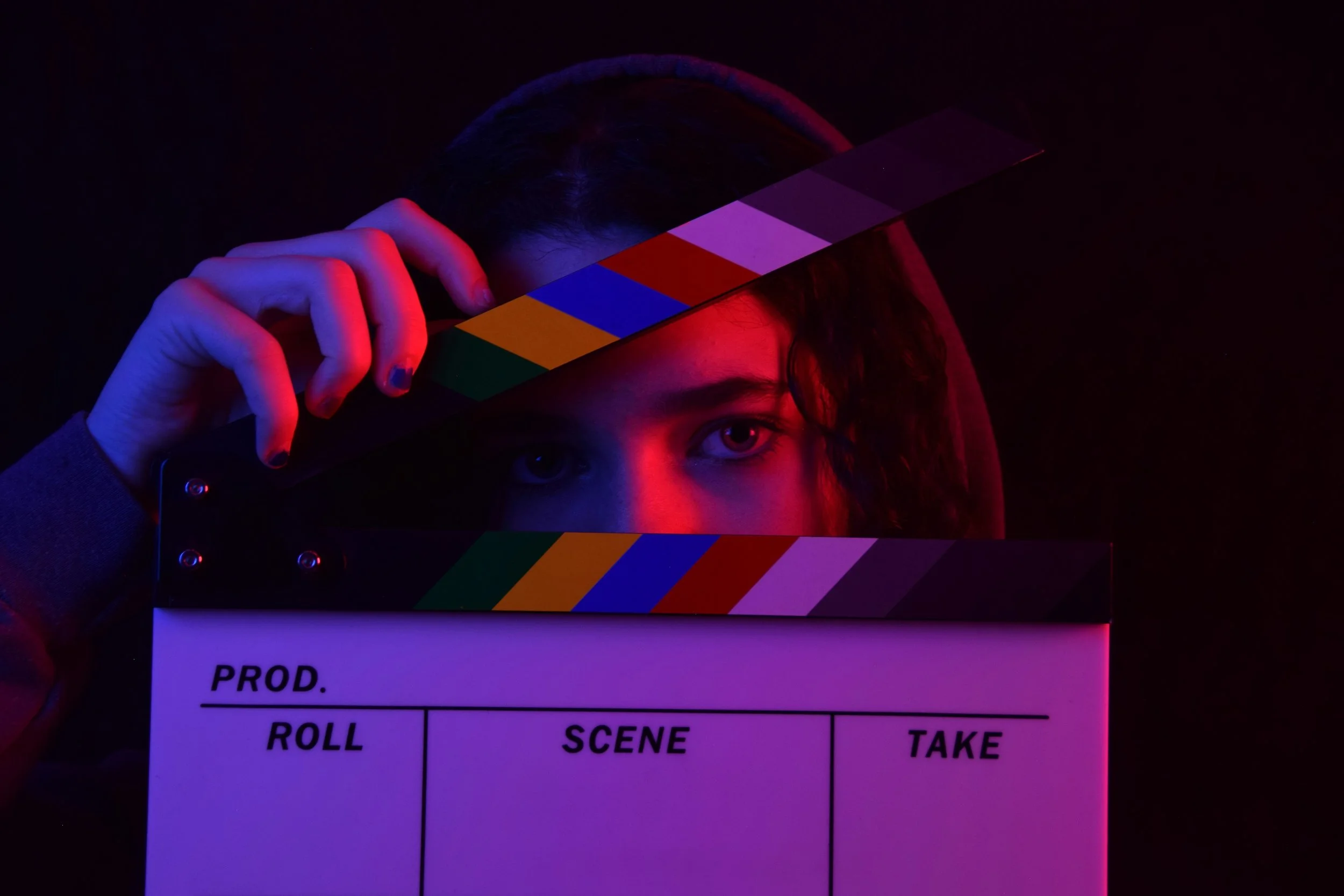 Person holding a film slate behind their face with blue, red, yellow, and purple lighting.