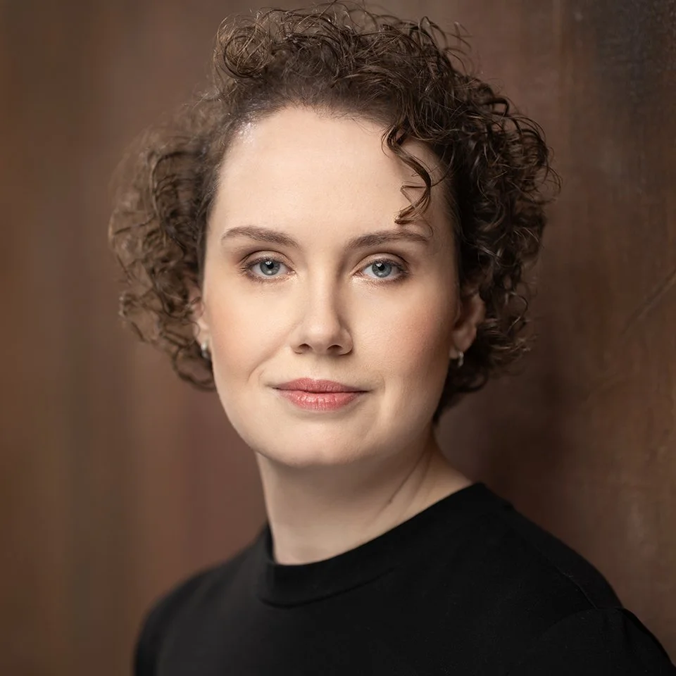 A photo of Julie Mallet-Paret, award winning director. Julie teaches improv, acting, voiceover, and business for performers. They have short, curly brown hair, blue eyes, and are wearing a black top, standing in front of a wooden background.