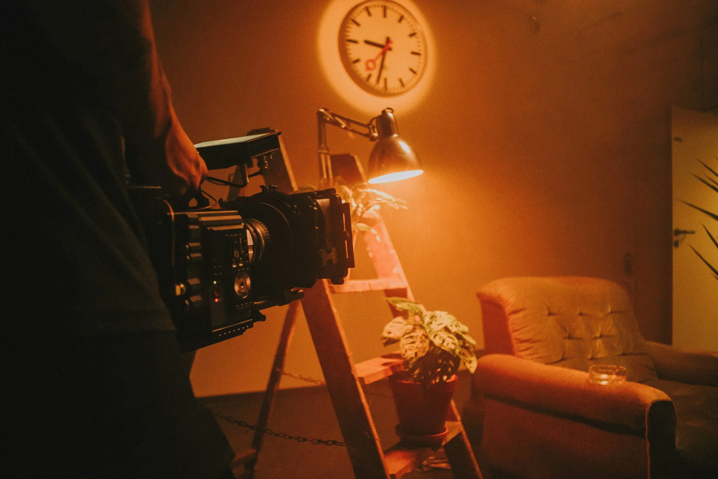 A person holding a camera, filming or taking photos indoors. The room has warm lighting, a wall clock showing 9:10, a potted plant, a wooden ladder shelf, an orange couch, and a desk lamp.