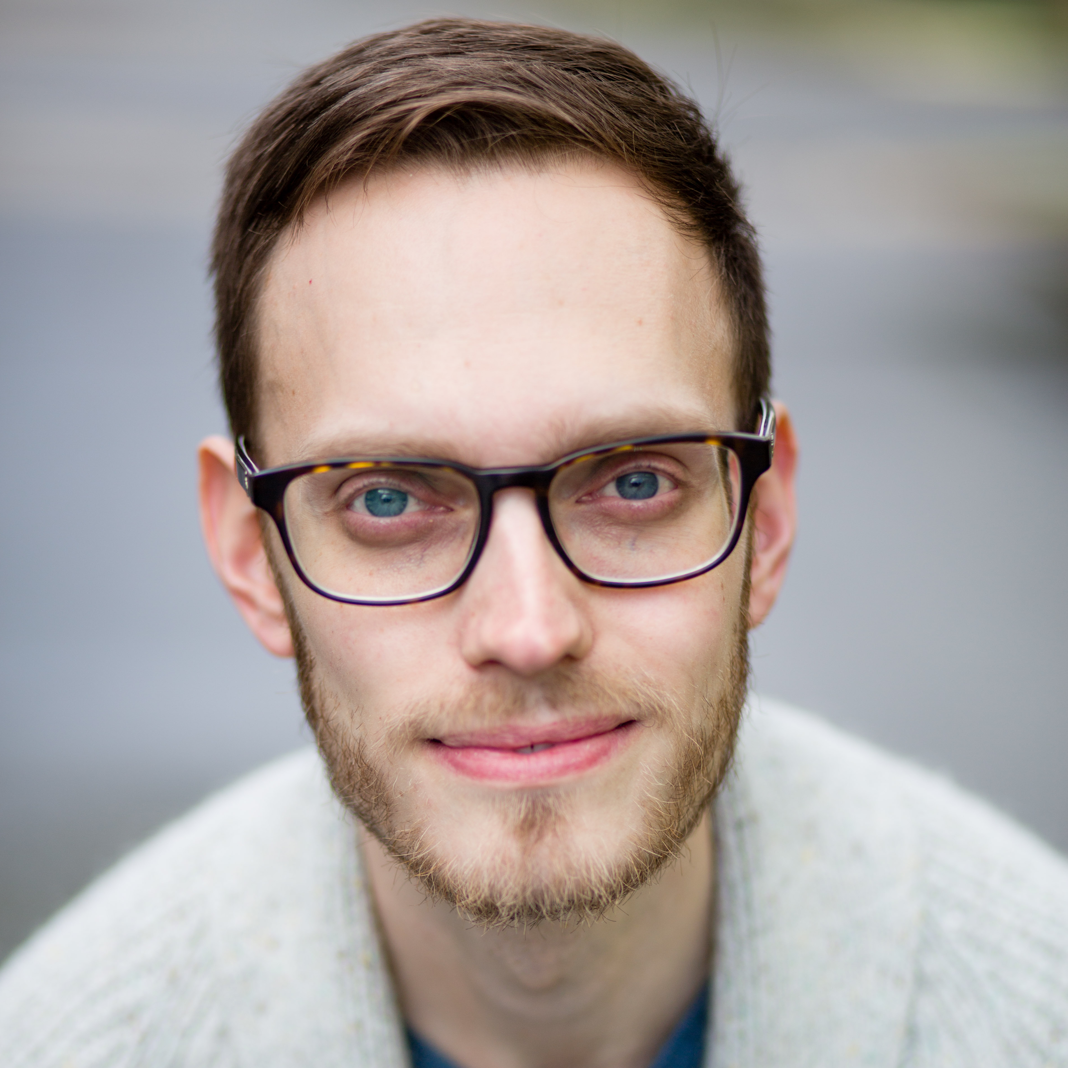 A photo of improv instructor Nikolai Witschl. He is a man with brown hair, glasses, blue eyes, and a light beard smiling outdoors.