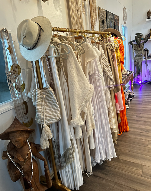 Clothing rack with white and beige dresses, a straw hat, and a white woven bag. Decorated room with mannequins in the background and purple lighting.