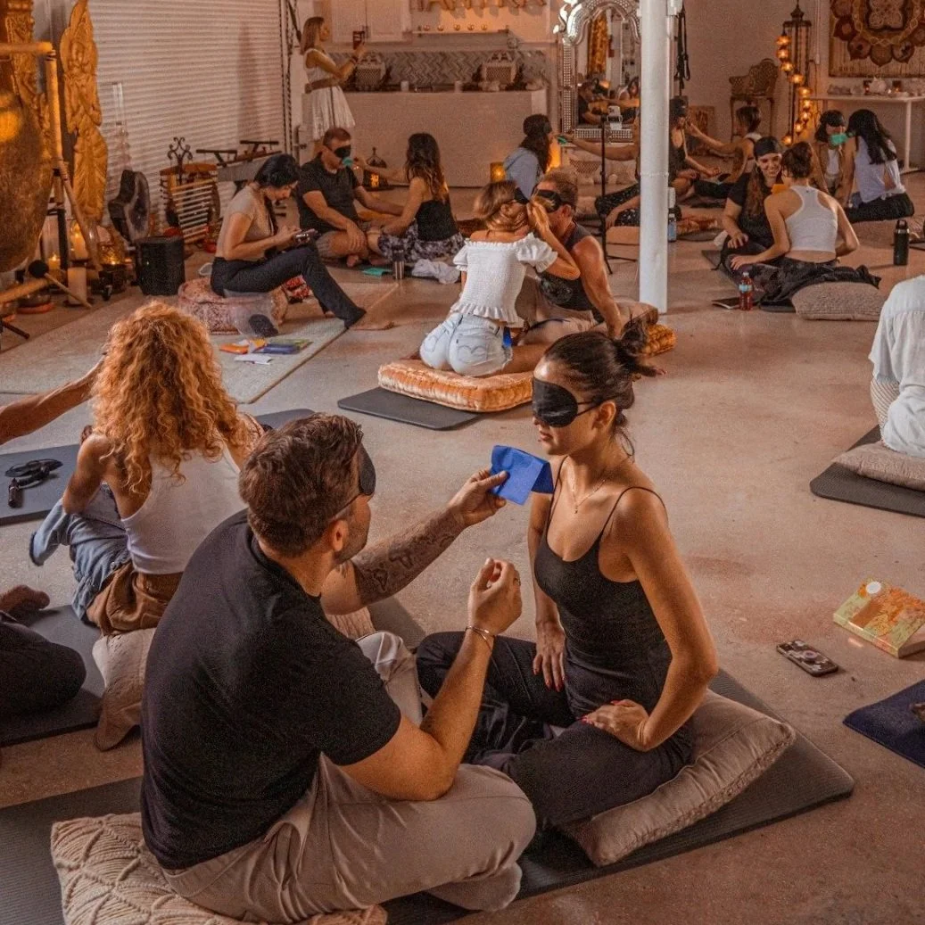 A group of people participating in an indoor sensory experience, with many wearing eye masks, seated on cushions and mats, in a warmly lit, decorated room.