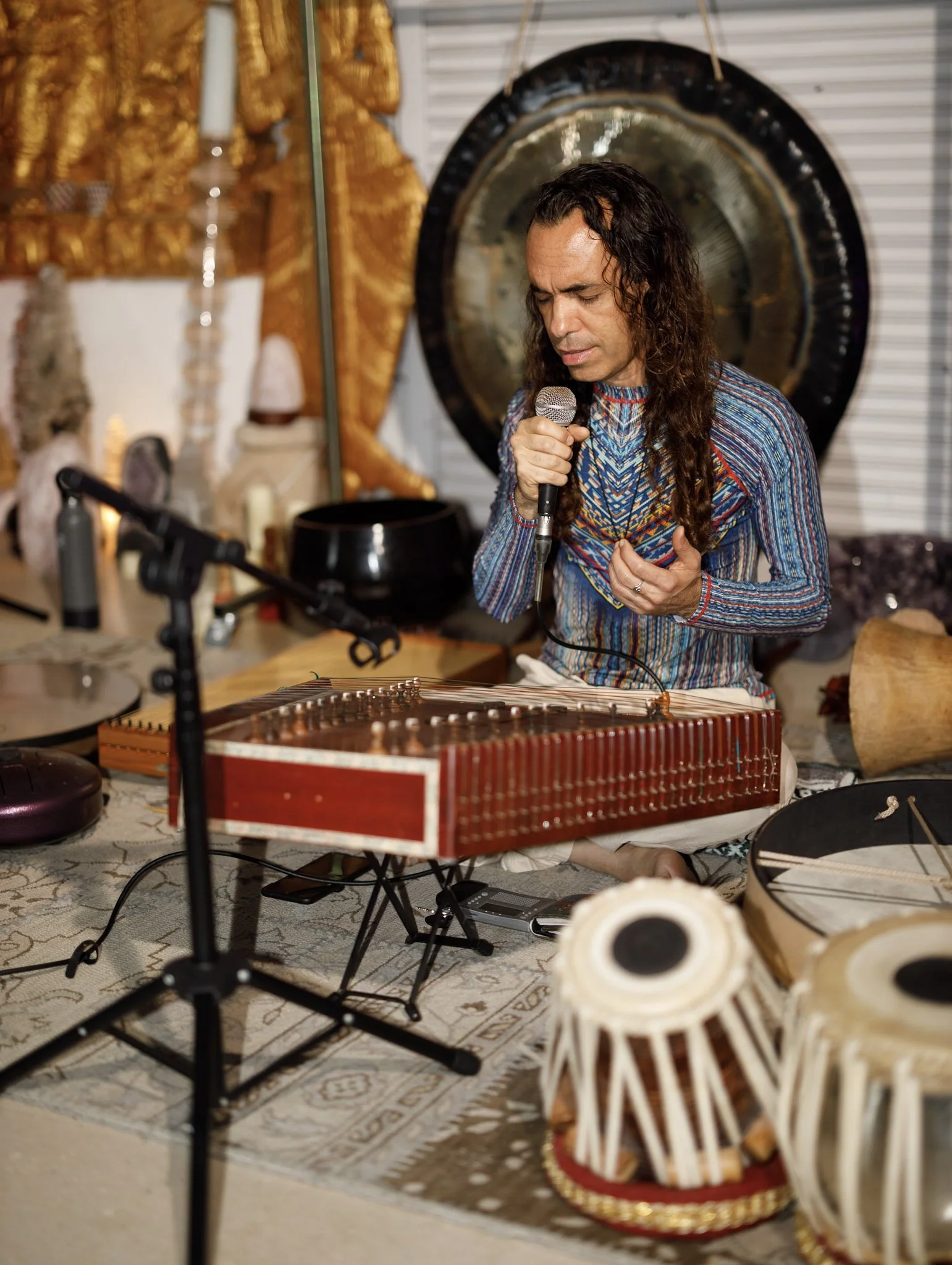 A man with long, curly hair and a colorful, patterned shirt plays a water gong with a mallet, surrounded by drums, a microphone, and musical instruments in a room decorated with gold and black artifacts and cultural items.