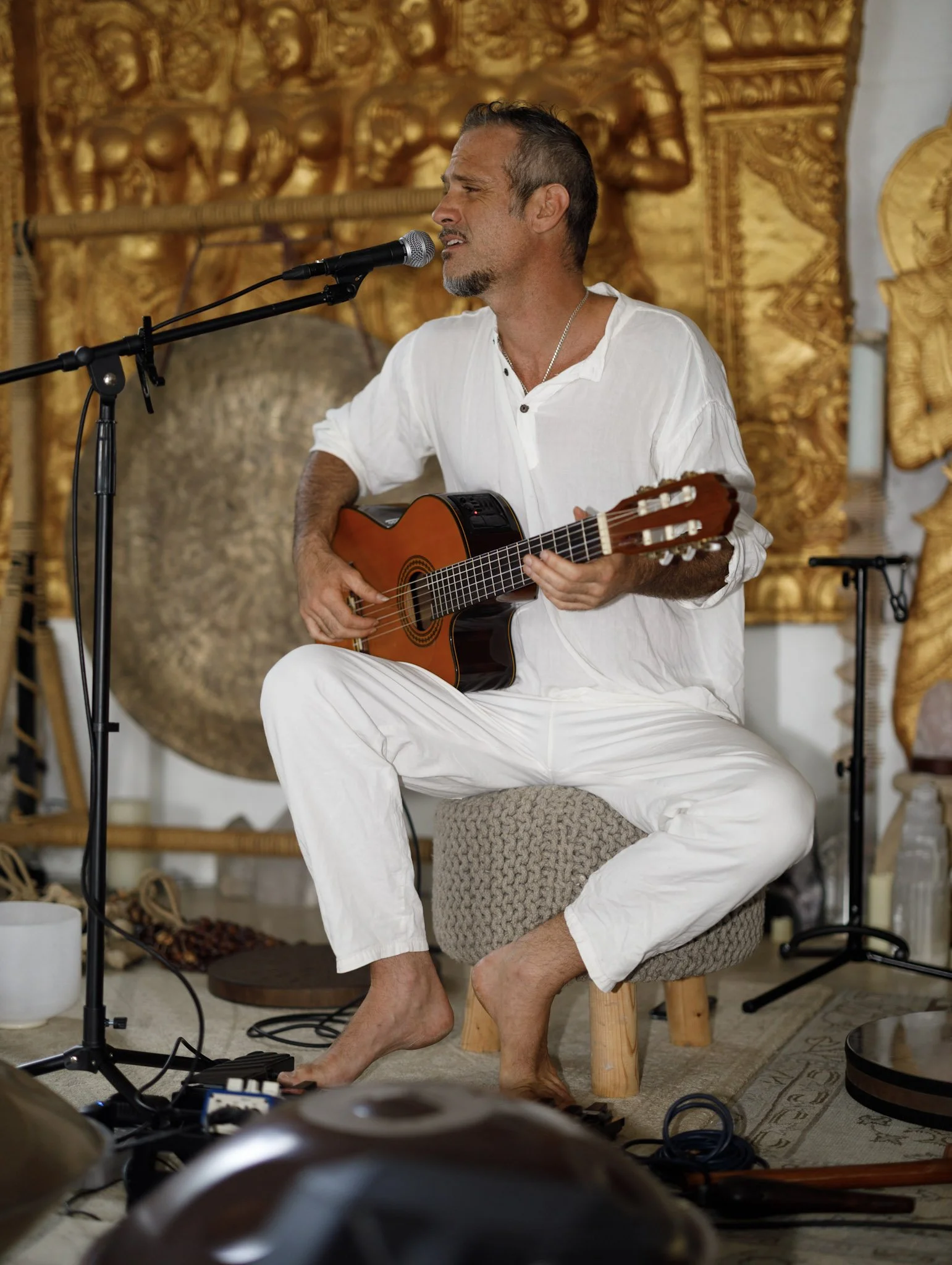 A man in white clothing sitting barefoot on a stool, playing an acoustic guitar and singing into a microphone, with a gold decorative wall in the background.