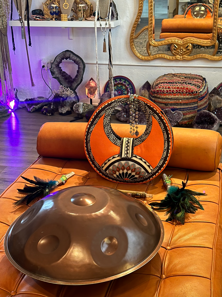 A round orange chair with a black and white bead embroidered drum-shaped bag resting on it. In front is a large metallic handpan drum with circular indentations. There are feathered accessories with beads on both sides of the drum. The background displays shelves with jewelry, crystals, and decorative items, and a mirror with a gold ornate frame hanging on the wall.