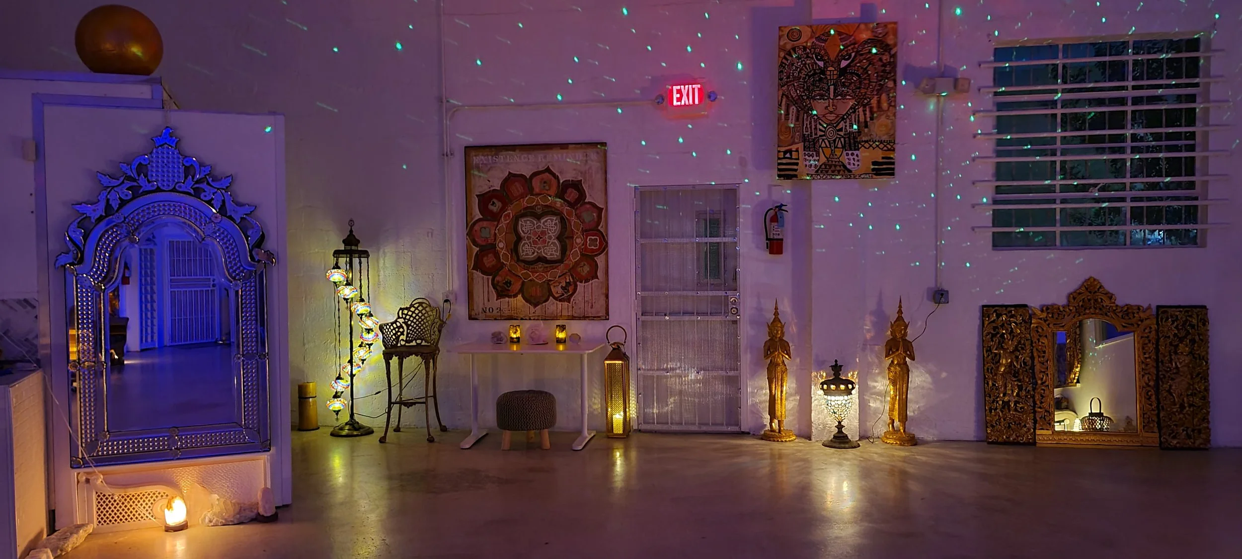An interior room decorated with Asian-inspired art and lighting. There is a large ornate mirror on the left, a string of colorful hanging lanterns, some small candles, and gold statues on the right. A wall painting and window are visible, along with 