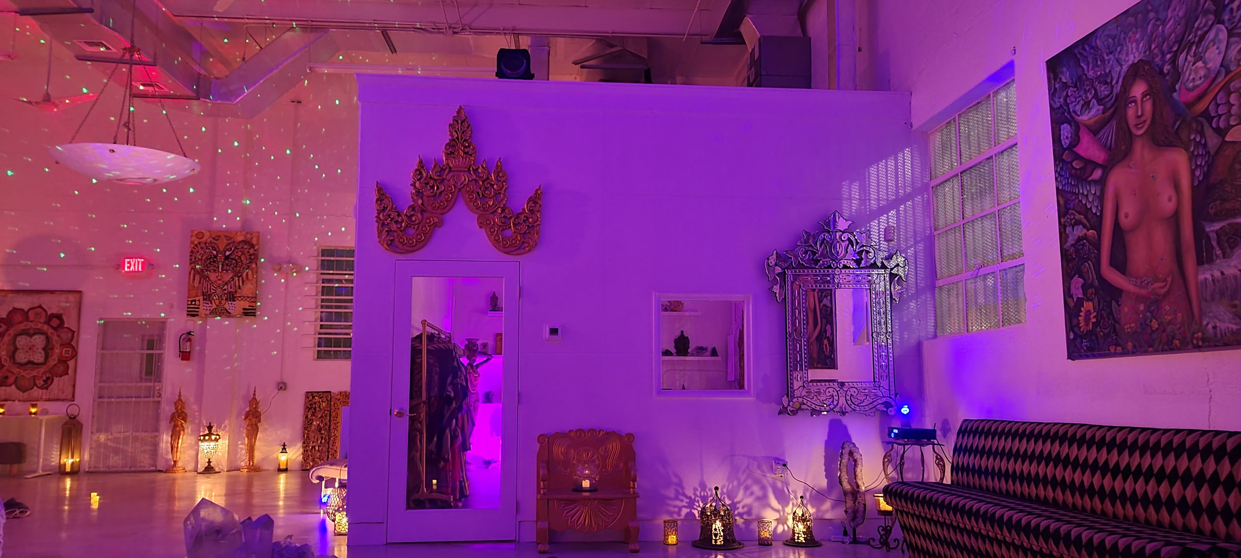 Interior of a room with purple and pink lighting, decorated with artwork, mirrors, and lanterns, featuring a patterned sofa and various wall art.