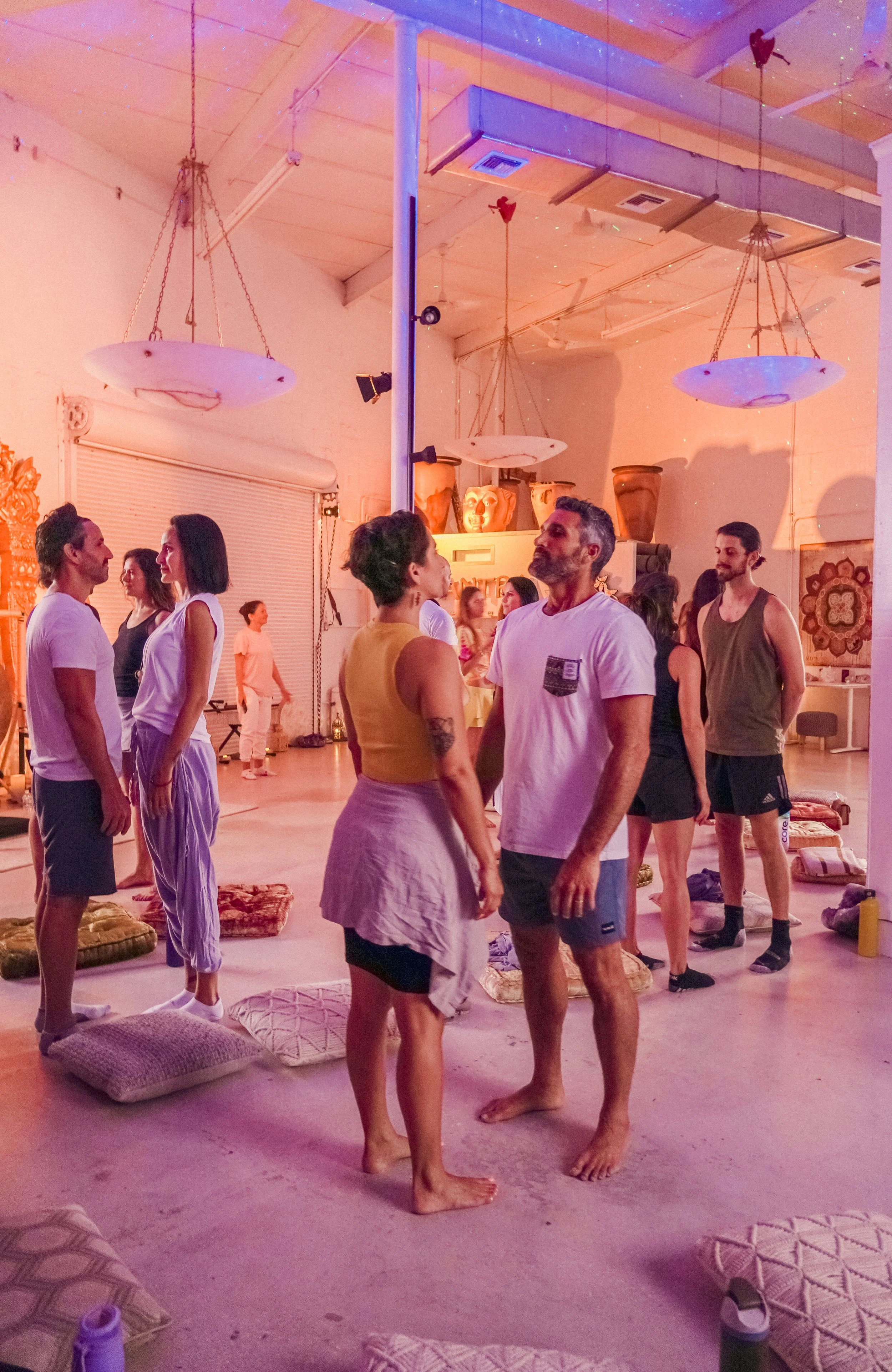 People participating in a meditation or yoga session inside a creatively decorated studio with hanging lights and large ceramic pots, sitting on cushions or standing in pairs.