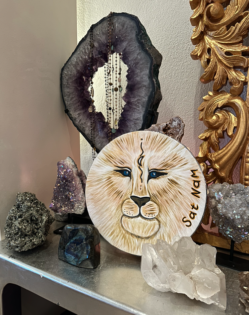 Decorative display featuring a painted lion face on a round surface, a large purple geode with jewelry hanging from it, smaller geodes and mineral specimens, and a gold ornate decorative piece in the background.