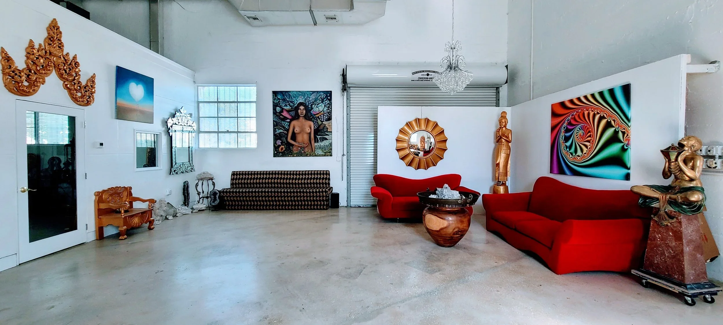 Interior of an art gallery with eclectic furnishings, including red sofas, a red armchair, a patterned black sofa, and a wooden bench. Decor includes artwork, sculptures, mirrors, and a chandelier.