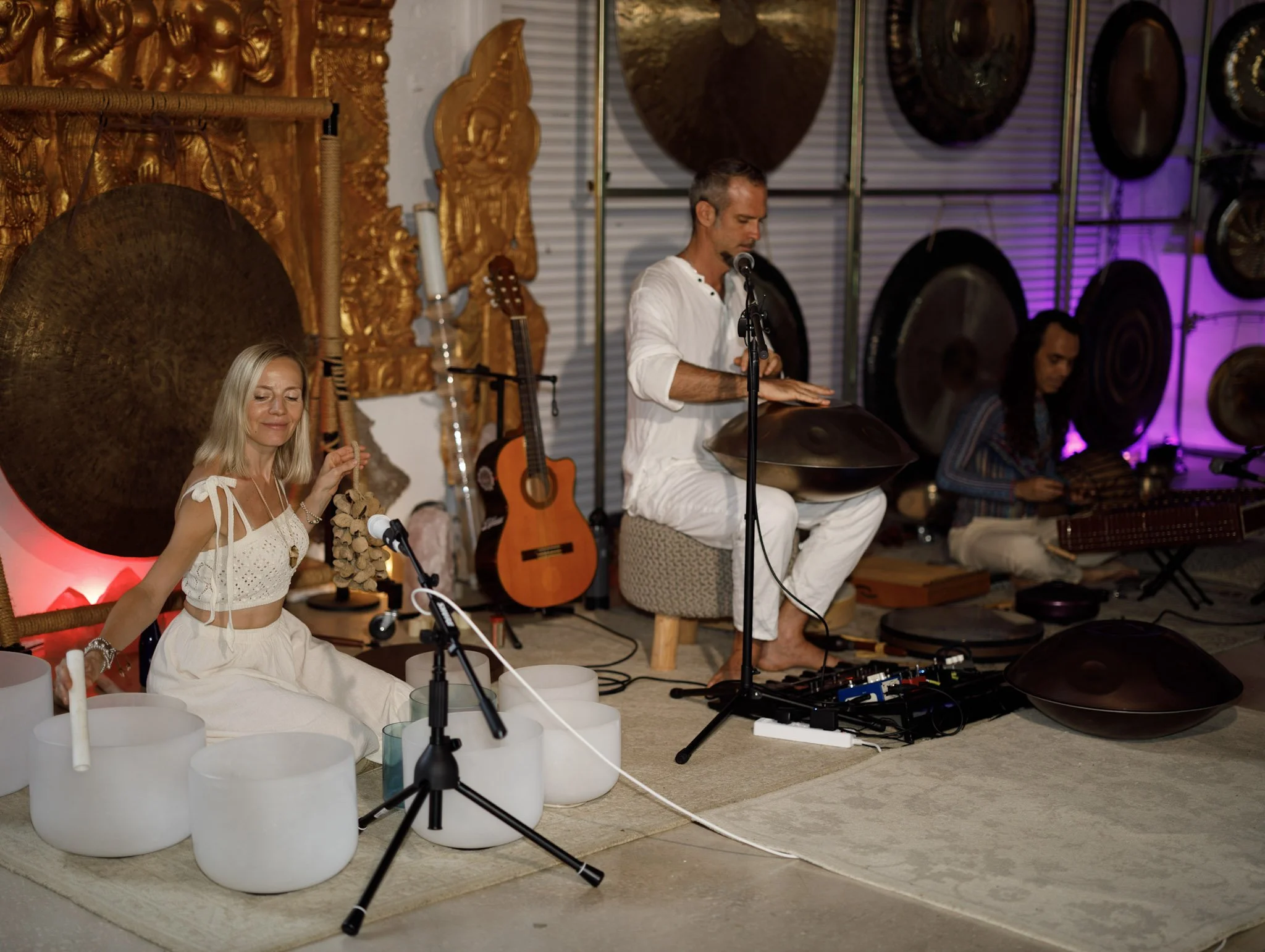 Musicians performing indoors with percussion instruments, guitars, and gongs, surrounded by ornate golden decorations and a purple-lit wall.