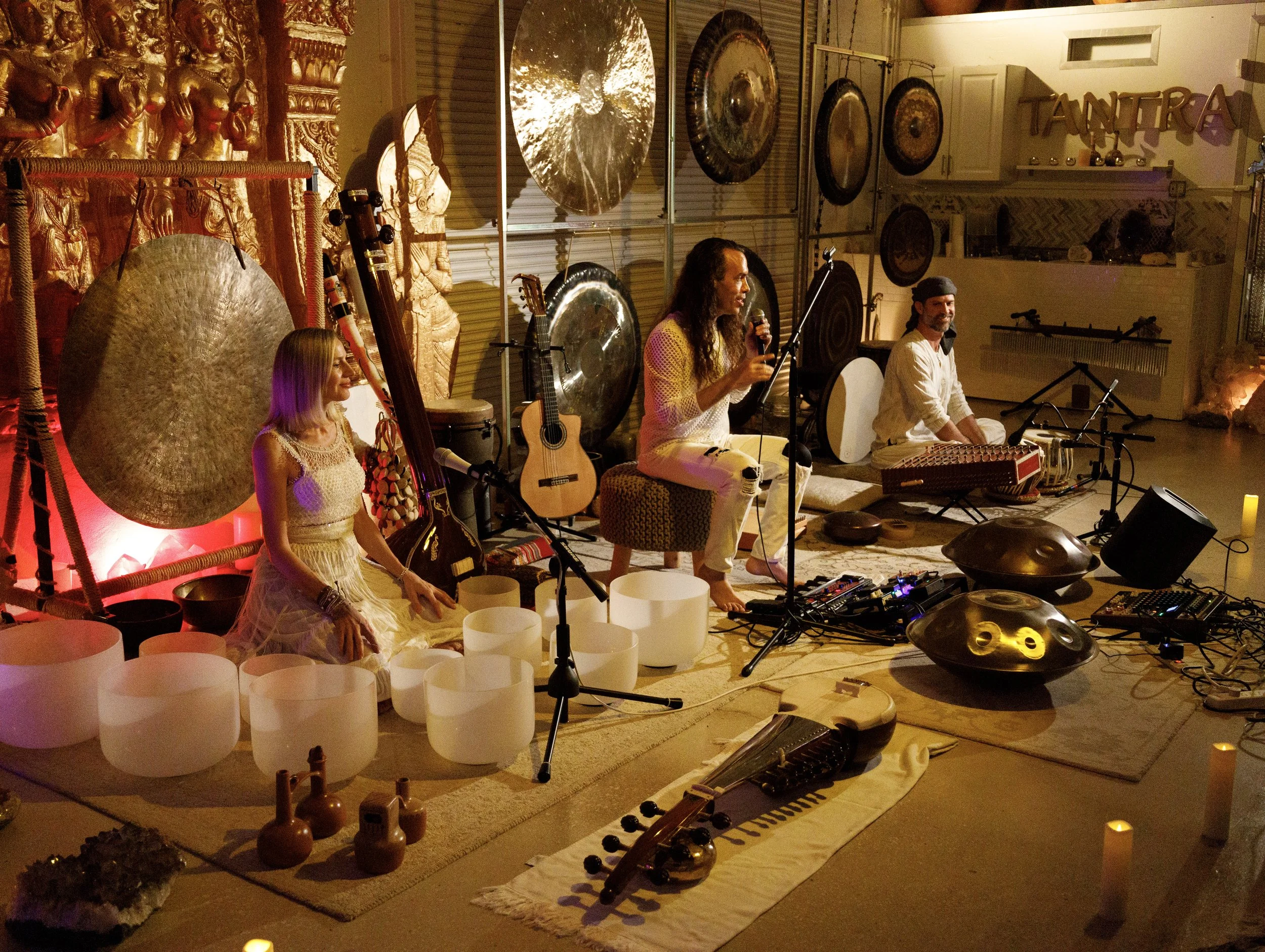 Musicians performing in a warmly lit indoor setting with various musical instruments and decorative elements, including gongs, guitars, and sculptures.