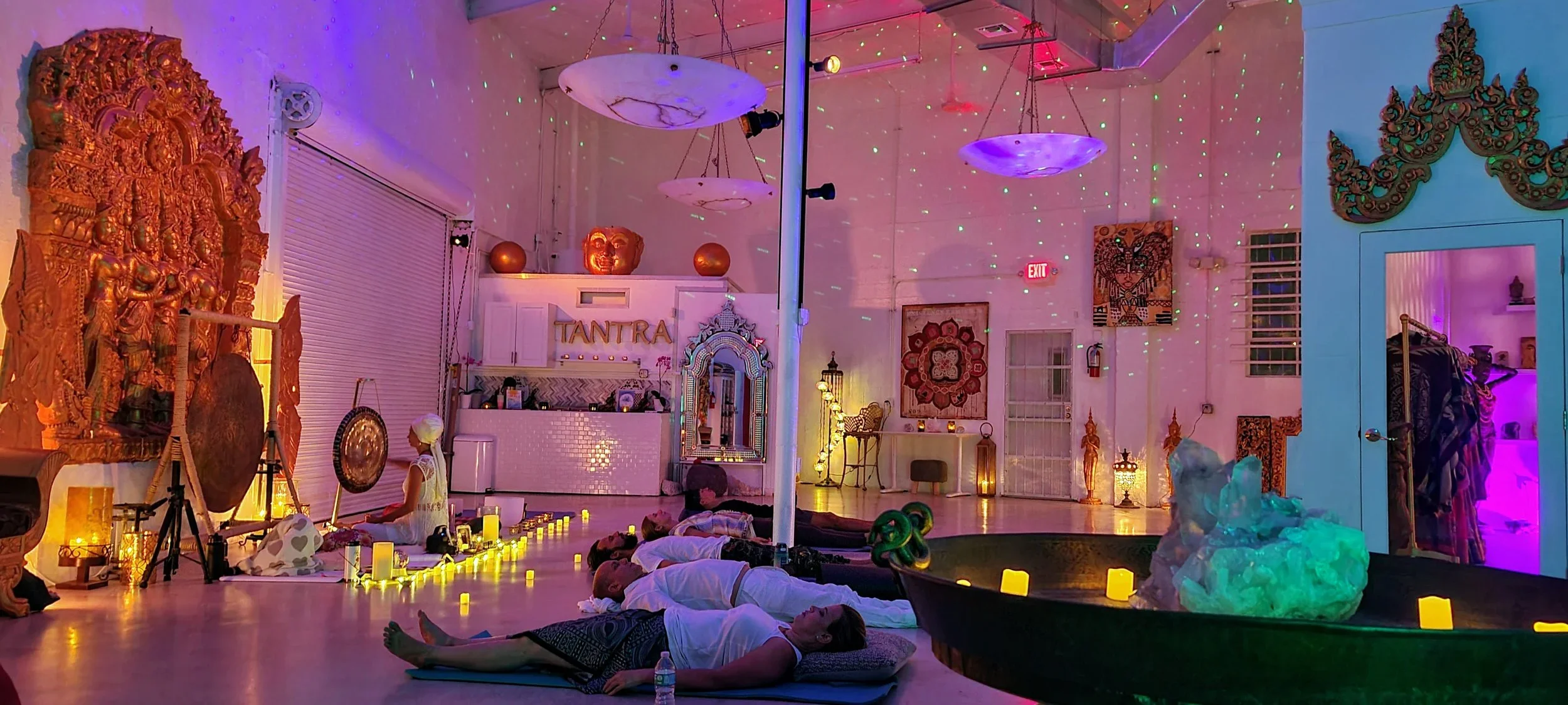 People practicing yoga or meditation in a dimly lit room decorated with Indian-inspired artwork and lighting, with candles and ambient lighting creating a calming atmosphere.