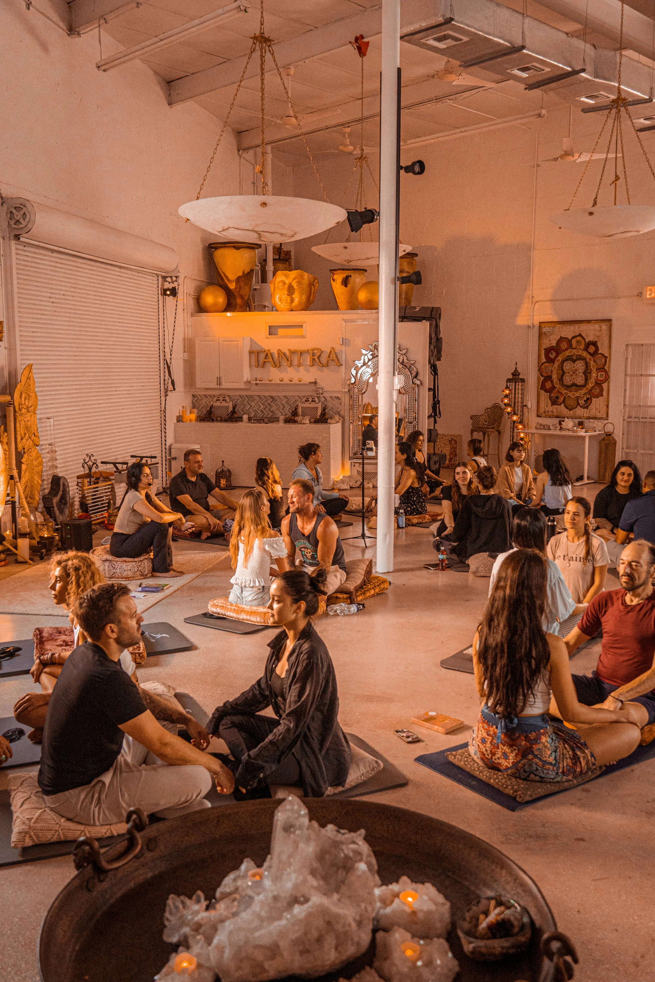 A group of people practicing yoga or meditation in a spacious, cozy indoor setting, seated on cushions and mats in pairs or groups, with warm, ambient lighting and decorative elements.