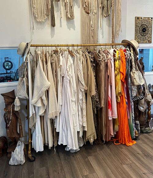 Clothing rack with beige, white, and colorful dresses and garments in a boutique store. Decor items and wall hangings in the background.