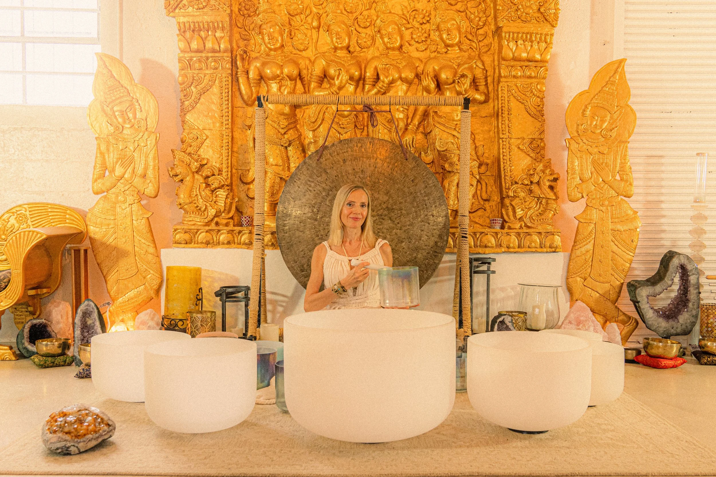 A woman with blonde hair, wearing a white dress, sitting in front of a large gong, holding a mallet. The background features a golden carved wall with figures and ornate decorations, and the setting is filled with crystals and spiritual objects.