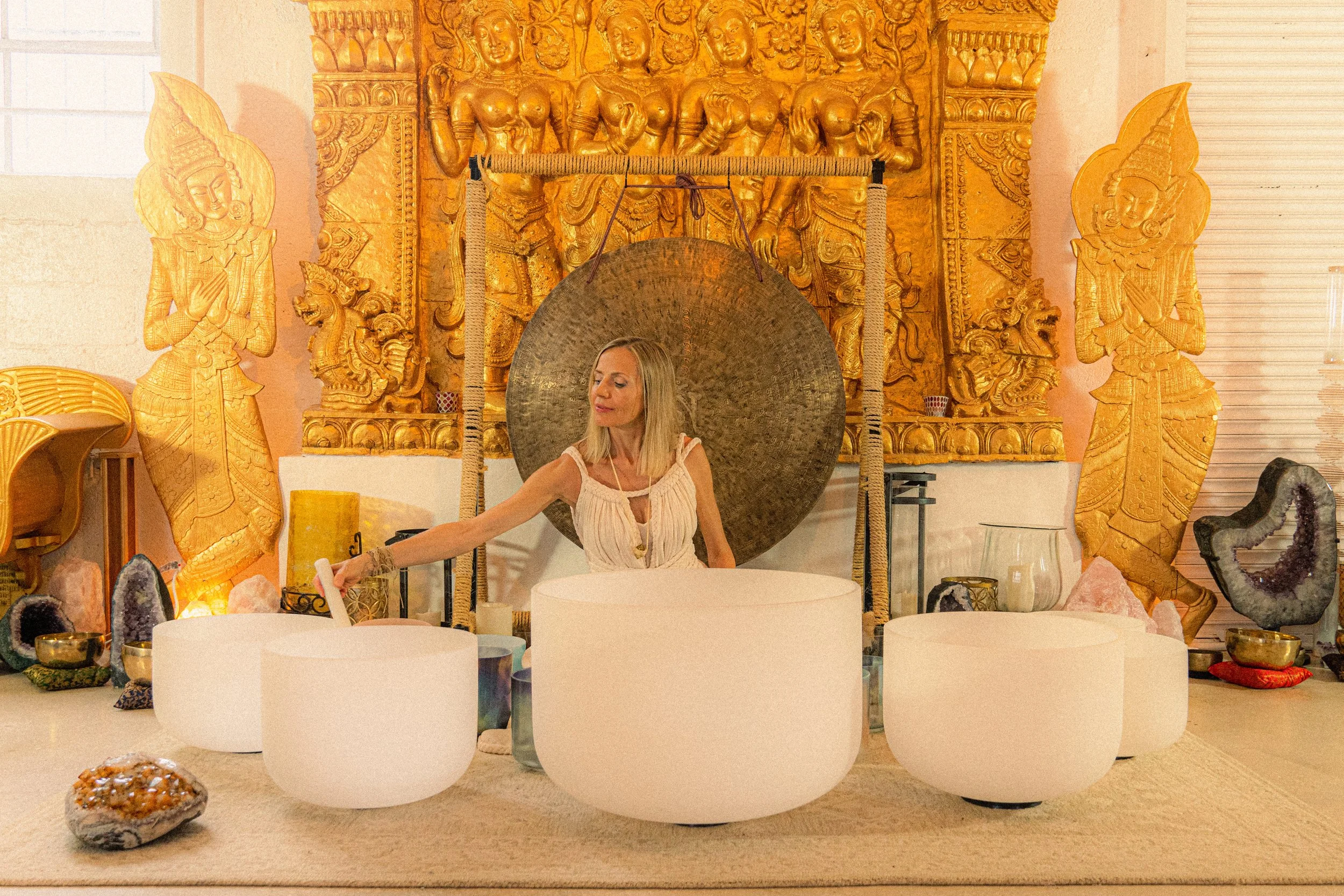 A woman playing crystal singing bowls in a room decorated with gold and crystal art pieces, including a large gong, sculptures, and geodes.