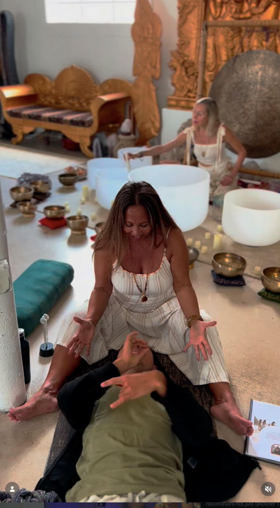 Two women are practicing sound healing with crystal bowls set on the floor, surrounded by candles, with one woman playing the bowls and the other sitting cross-legged, engaging in the experience.