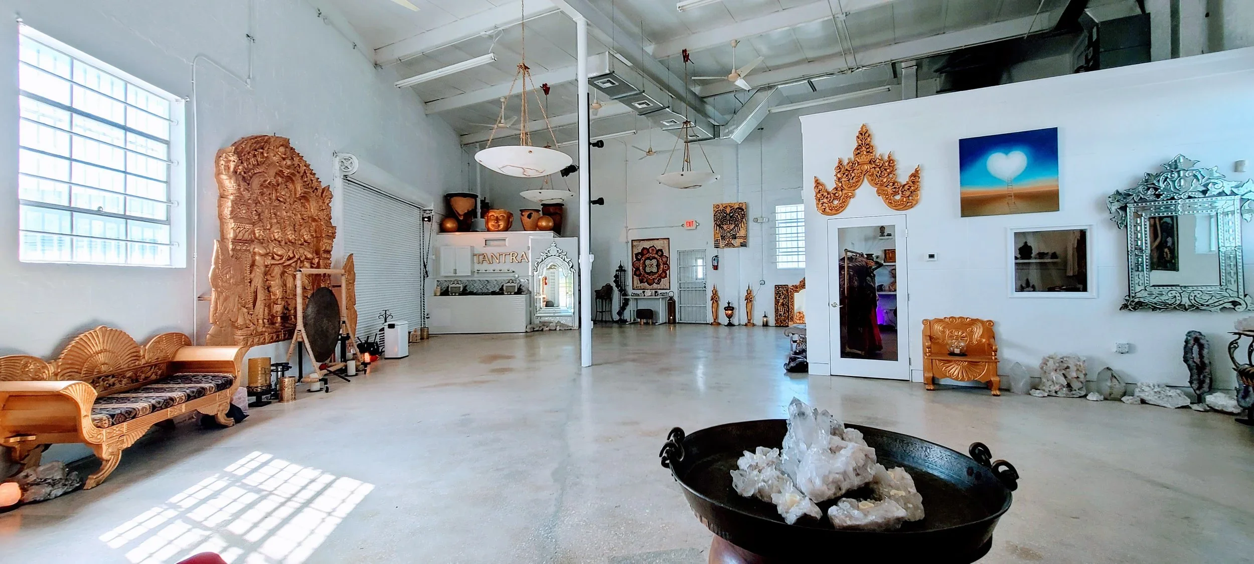An interior space decorated with various Asian-inspired art and artifacts, including a large wooden carving, ornate mirrors, and sculptures, with natural light coming through the windows.