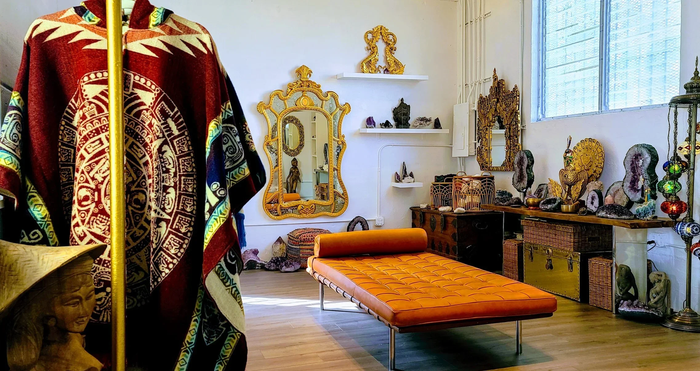 An interior room decorated with various cultural artifacts. In the foreground, there is a colorful, patterned cloak hanging on a yellow pole. The room features a large, orange cushioned lounge chair, a gold ornate mirror, and a wooden table with decorative rocks, sculptures, and mosaics. There are on-wall and shelf displays of sculptures, framed mirrors, and crystals, along with wicker storage chests and a tall standing lamp with hanging colorful ornament spheres. The room has white walls, a large window with metal bars, and wooden floors.