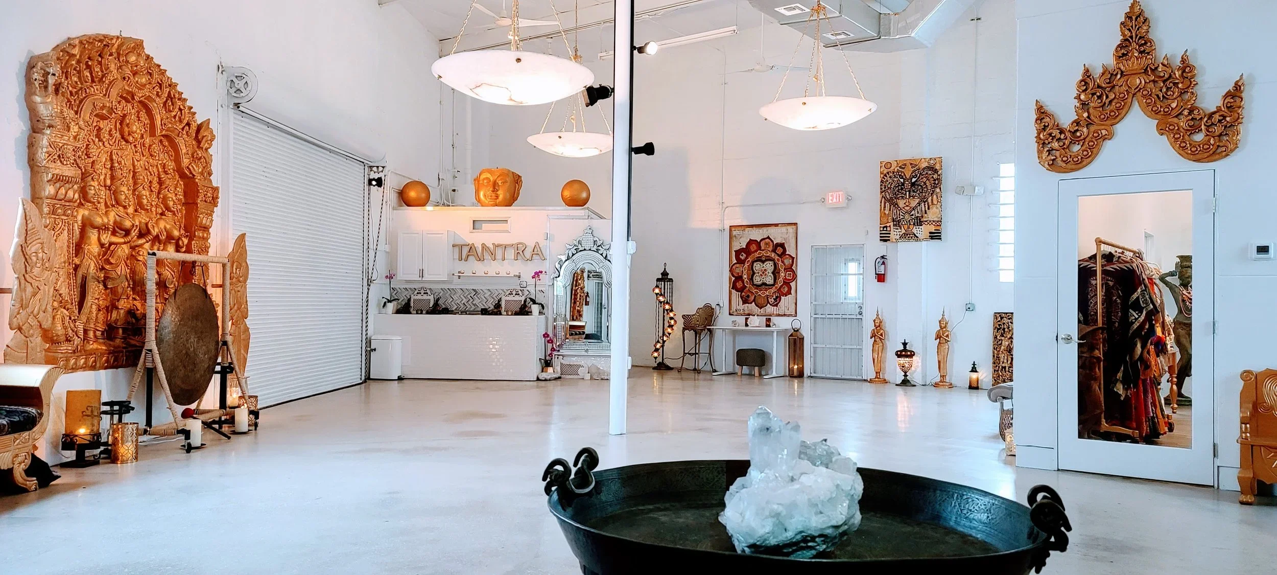 Spacious wellness or yoga studio with white walls and diverse Indian-inspired decor, including carved wooden sculptures, wall hangings, and statues, with soft candle lighting and a reception area labeled 'TANTRA'