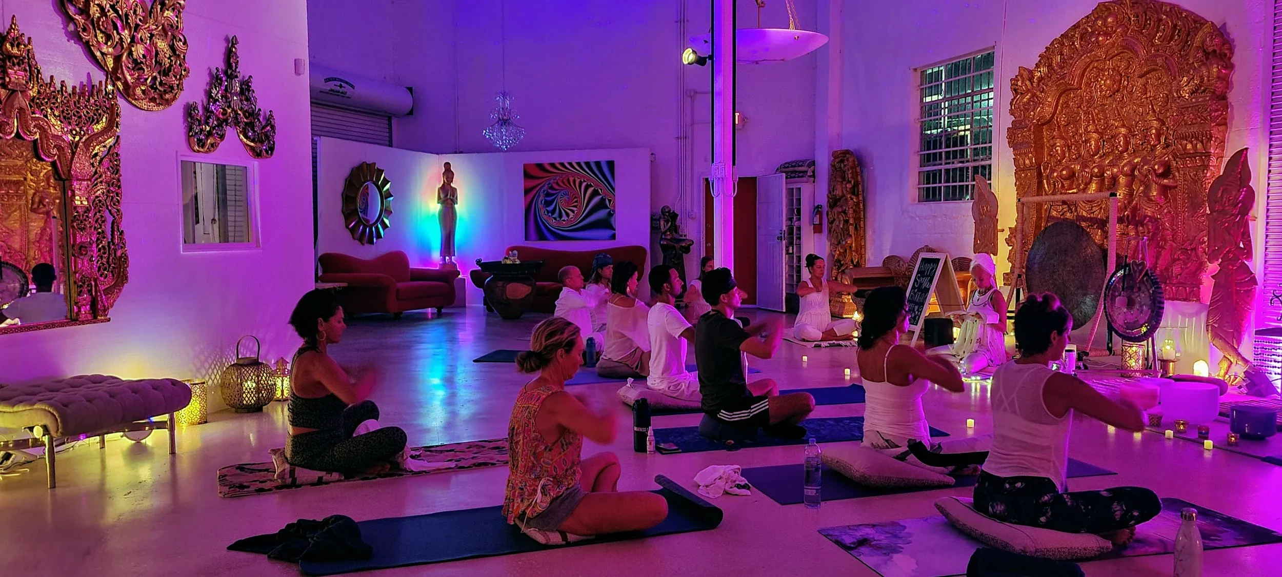 People participating in a yoga or meditation class in a room with colorful, vibrant lighting and ornate decorations, including sculptures and paintings, creating a spiritual and relaxing atmosphere.