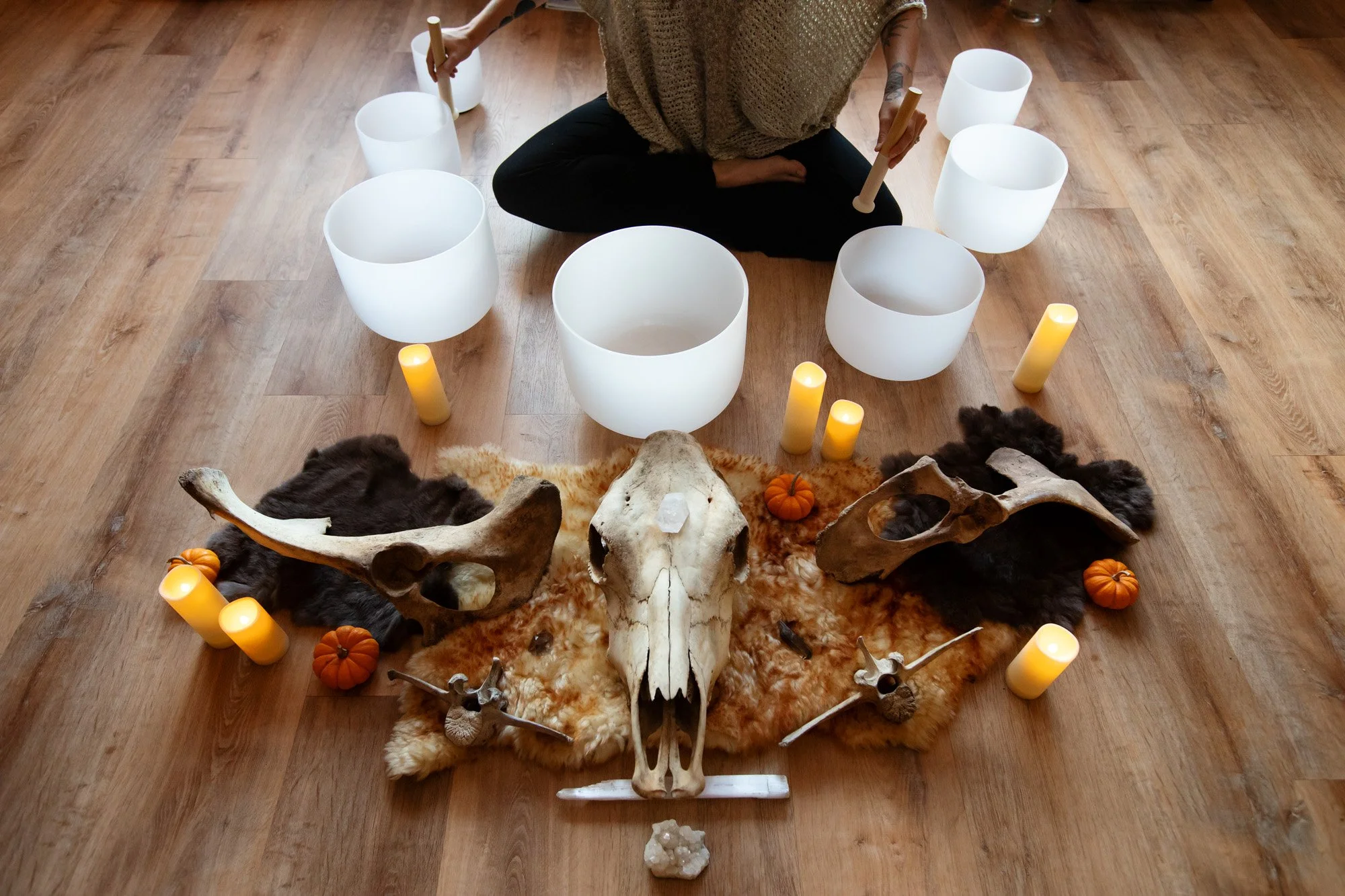 A person seated on a wooden floor surrounded by white crystal singing bowls, candles, small pumpkins, and animal skulls and bones, with a fuzzy fur rug underneath.