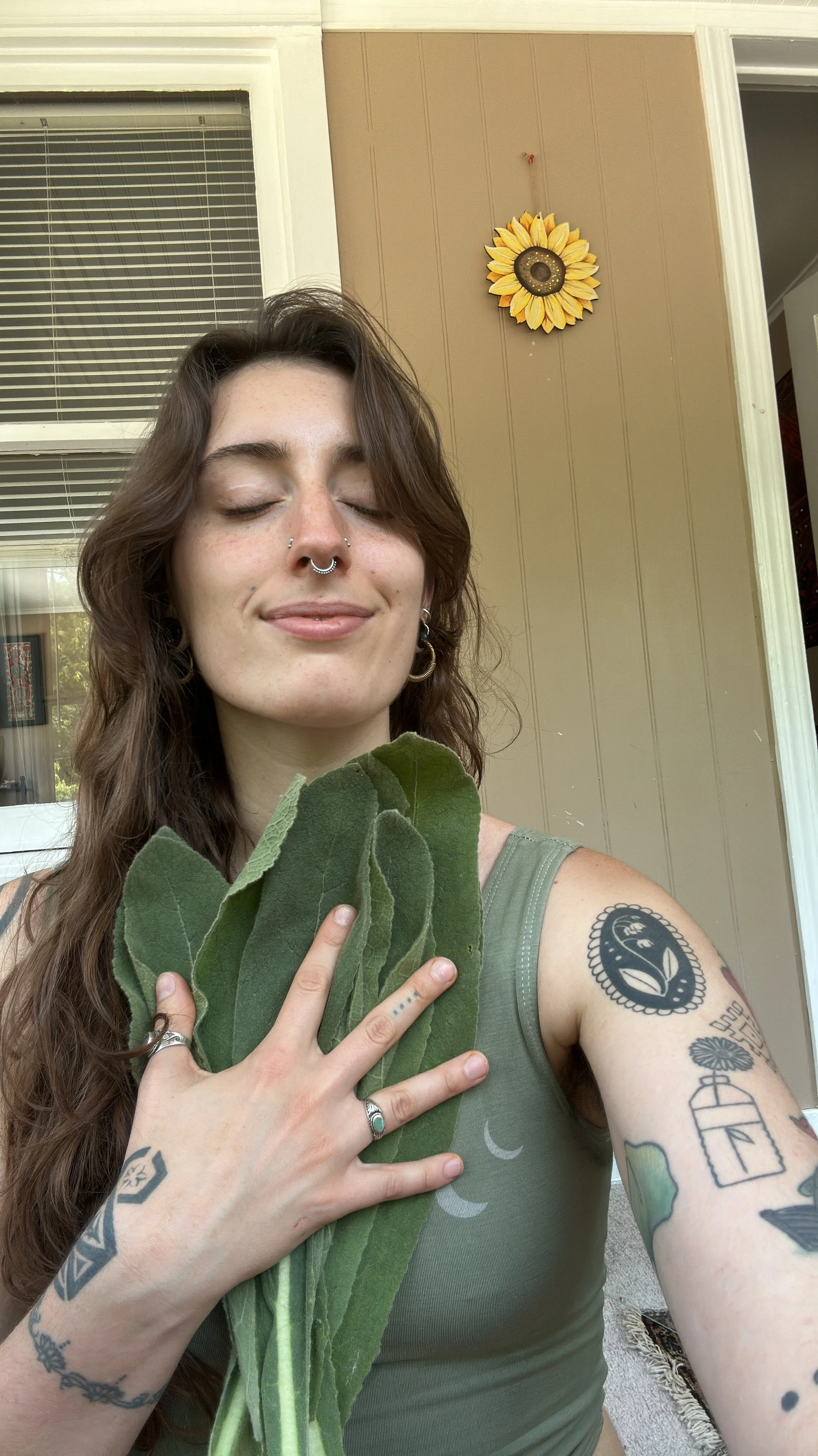 A smiling woman with long wavy brown hair, multiple tattoos on her arms, wearing rings, holding green leaves against her chest, standing outside on a porch with a sunflower decoration on the wall behind her.