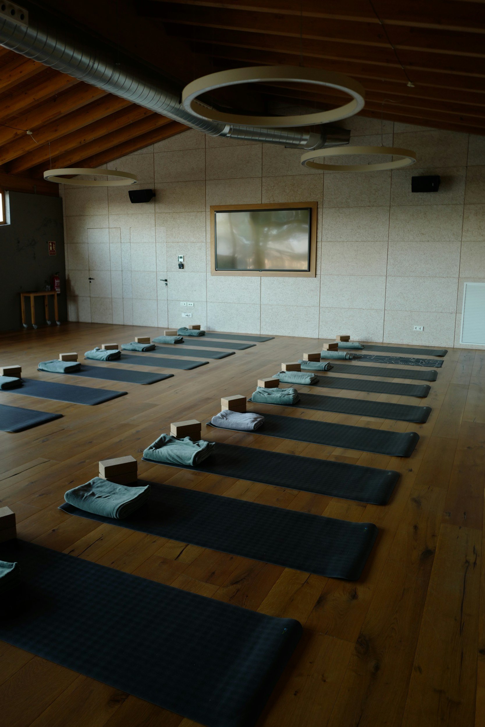 Yoga studio with black mats, folded towels, and wooden blocks arranged in a grid, a large monitor on the wall, and modern circular light fixtures hanging from a wooden ceiling.