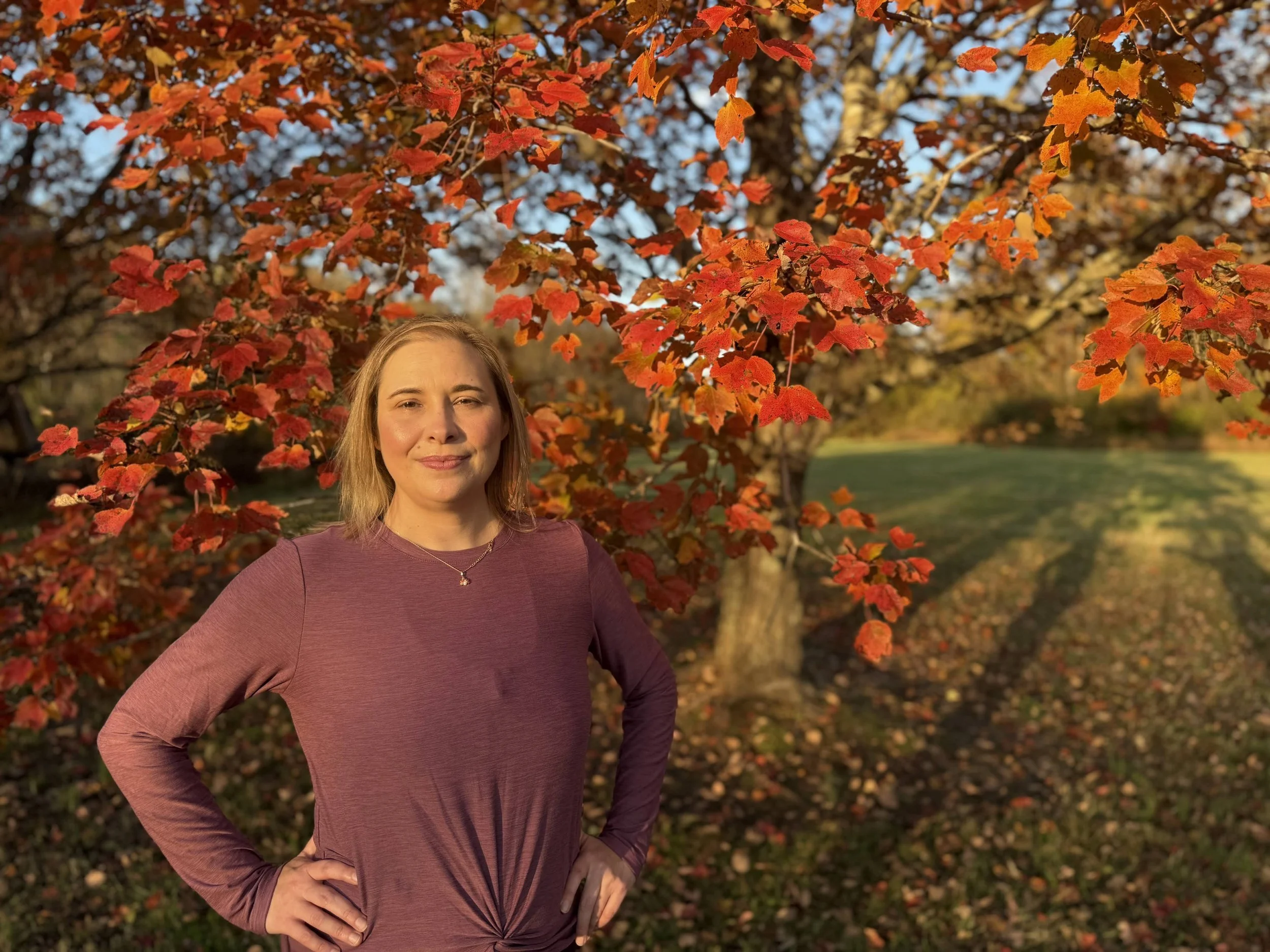 A woman standing outdoors in fall, posing with hands on hips in front of a large tree with orange and red autumn leaves, during late afternoon sunlight.