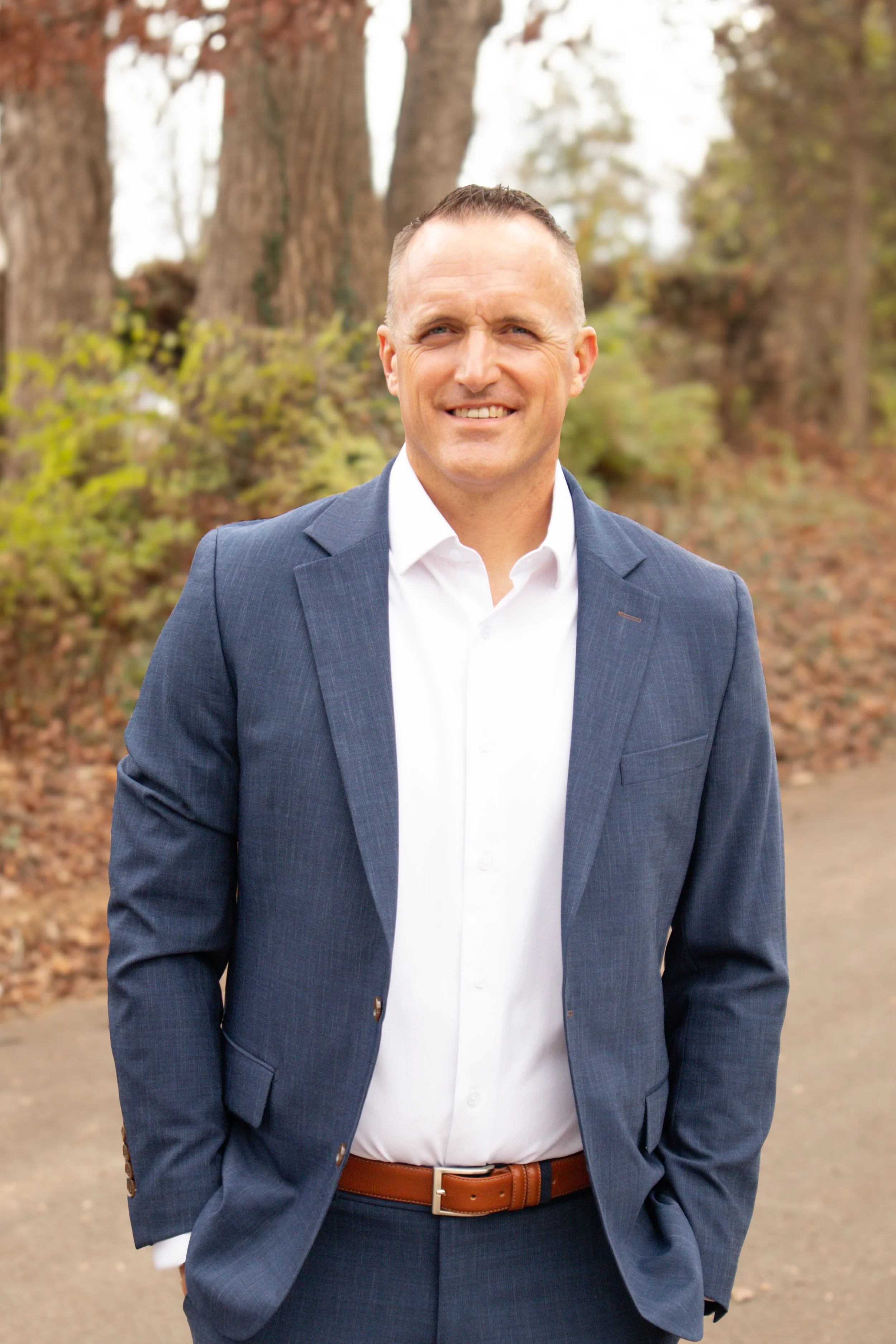 A man in a blue suit and white shirt standing outdoors on a dirt path with trees and leaves in the background.