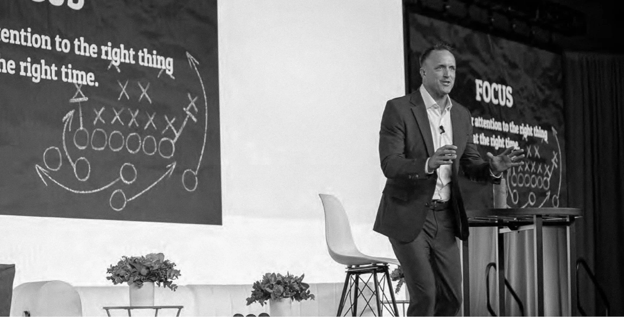 A man in a suit giving a presentation on stage with a large screen behind him displaying diagram and text about focus, with a small table and flower arrangements in the foreground.
