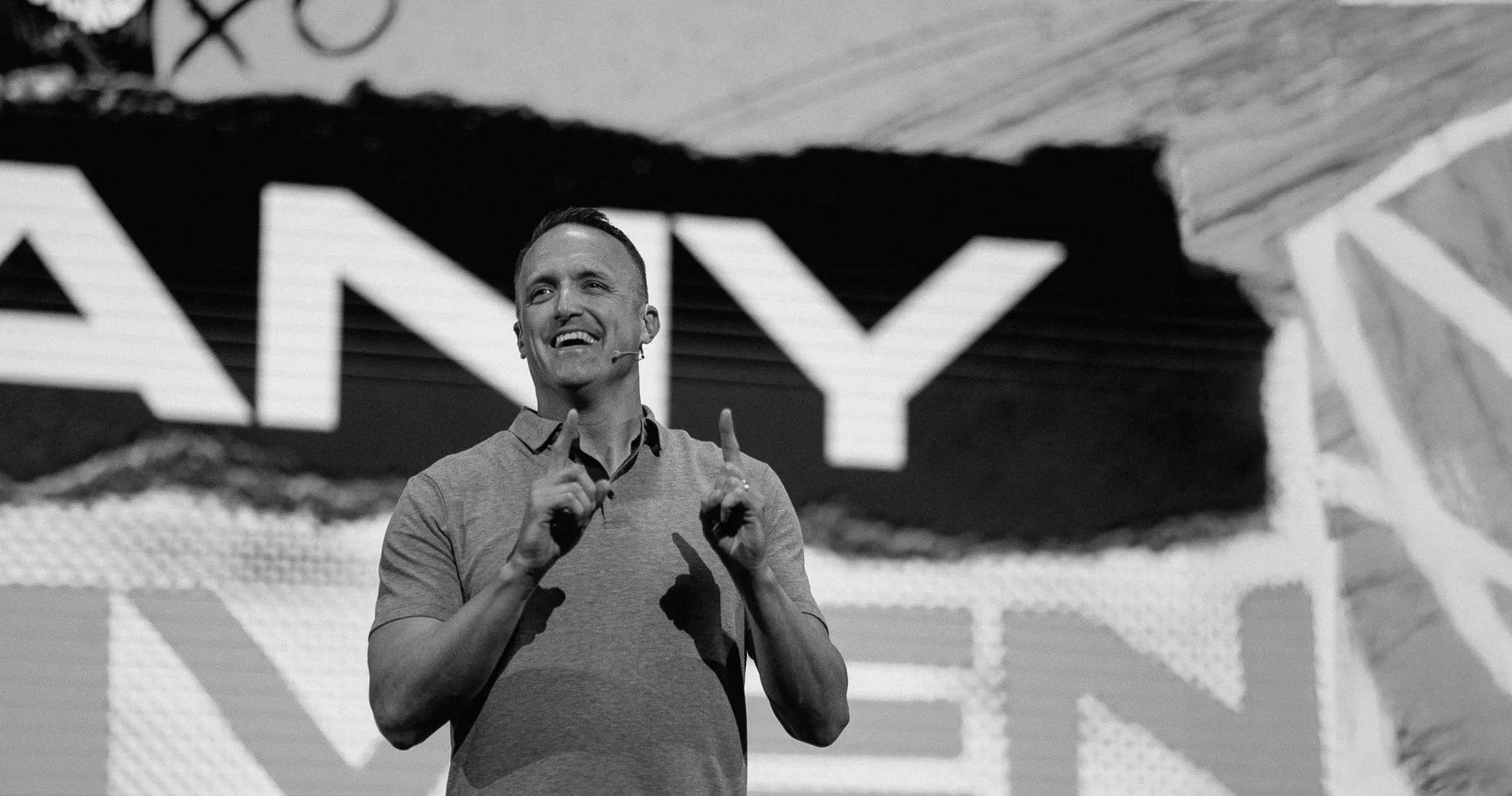 A man speaking on stage with a microphone headset, smiling, wearing a polo shirt, in front of a large screen