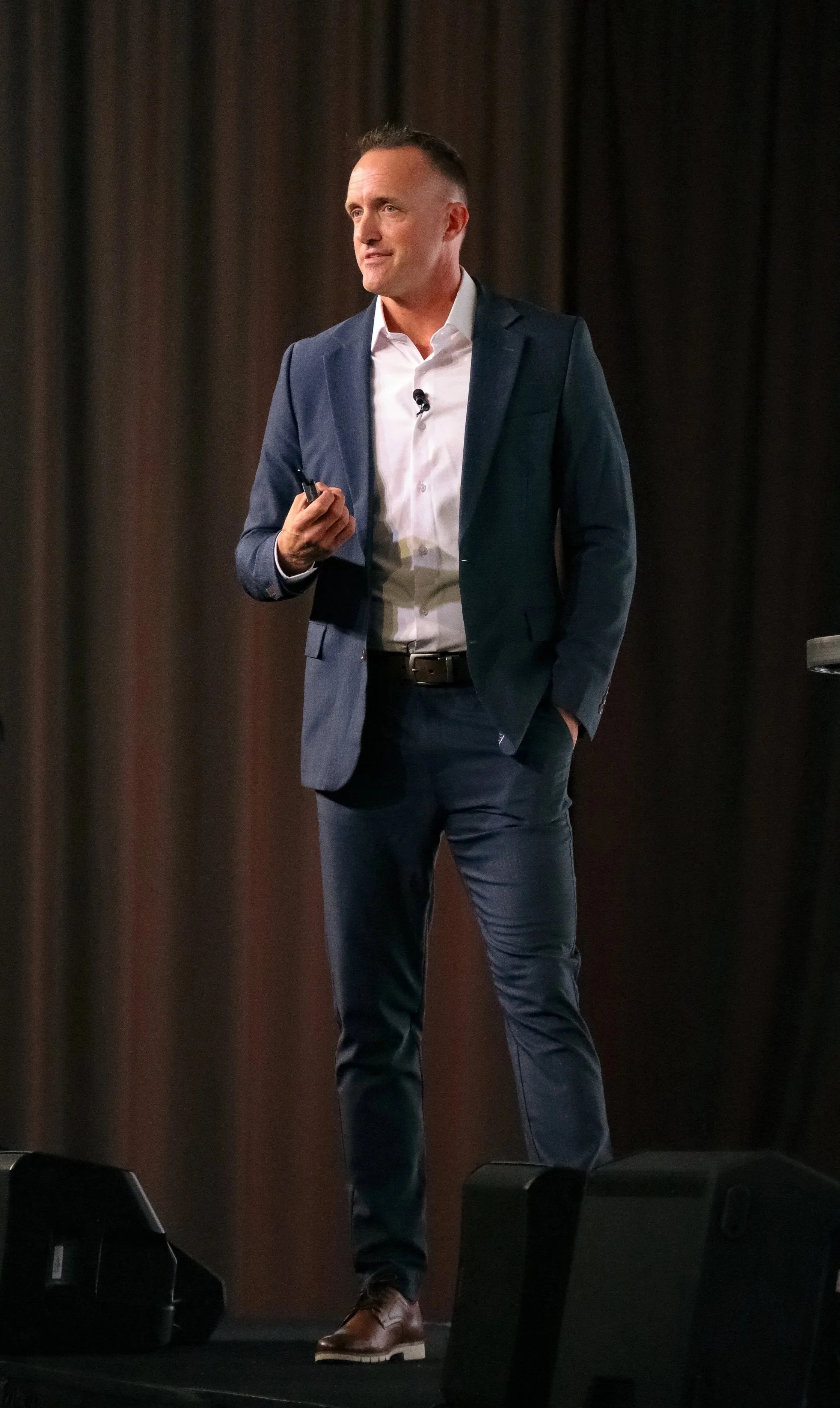 A man in a dark suit and white shirt is standing on stage in front of a dark curtain, holding a microphone and a clicker.