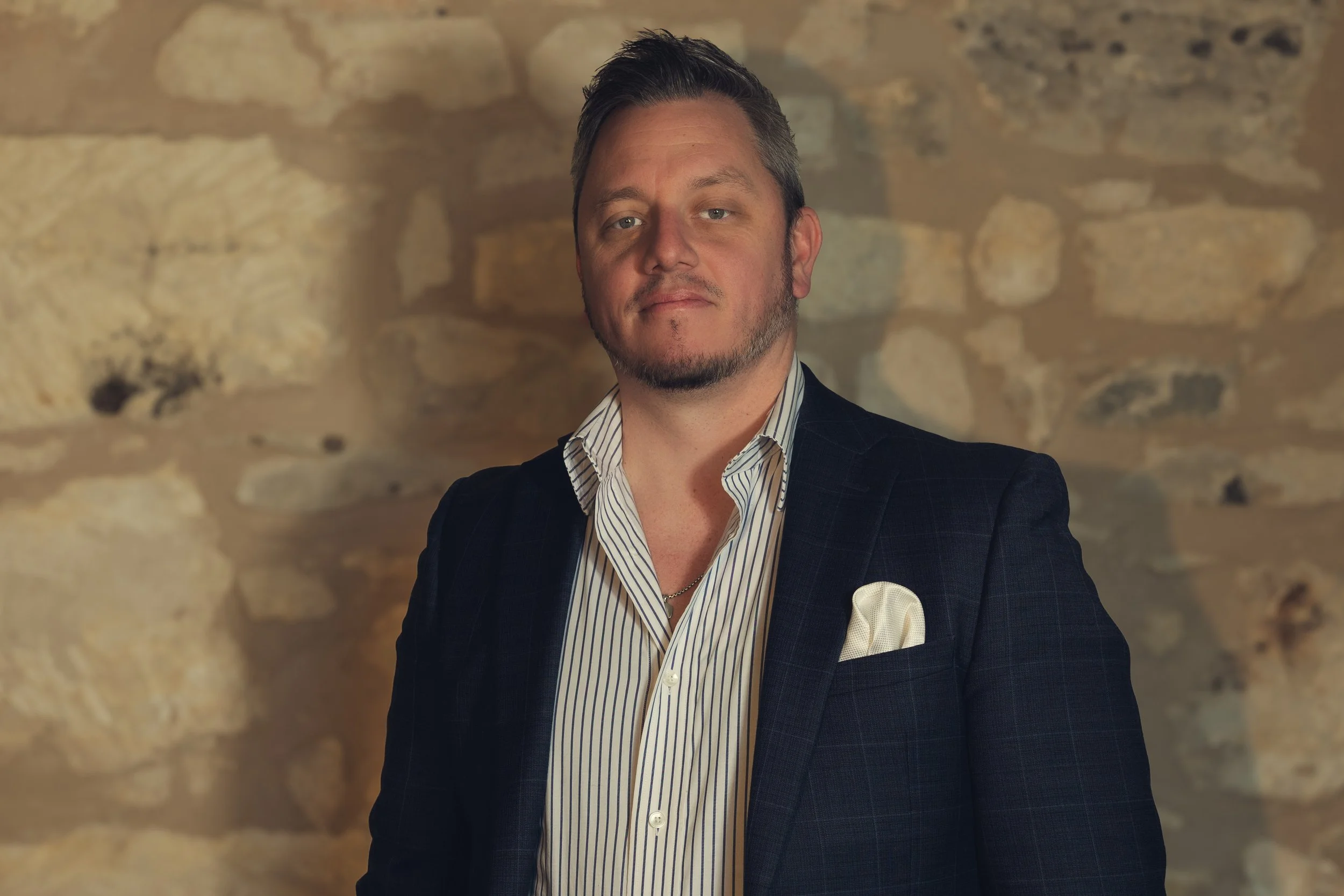 A man with short dark hair and a goatee stands in front of a stone wall, wearing a dark blazer, a striped shirt, and a white pocket square.