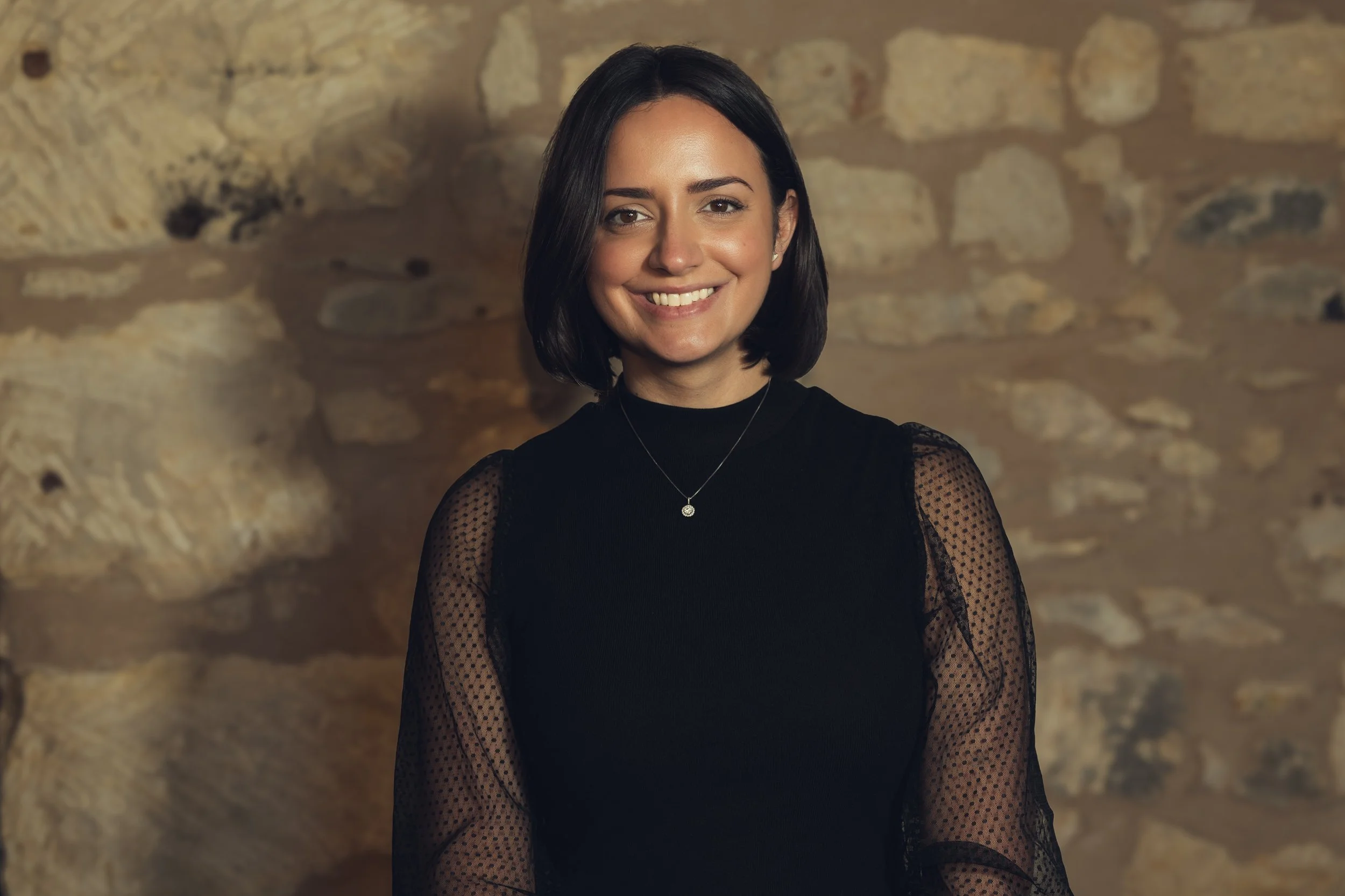 A woman with shoulder-length dark hair, wearing a black top with sheer, polka-dotted sleeves, smiling in front of a stone wall.