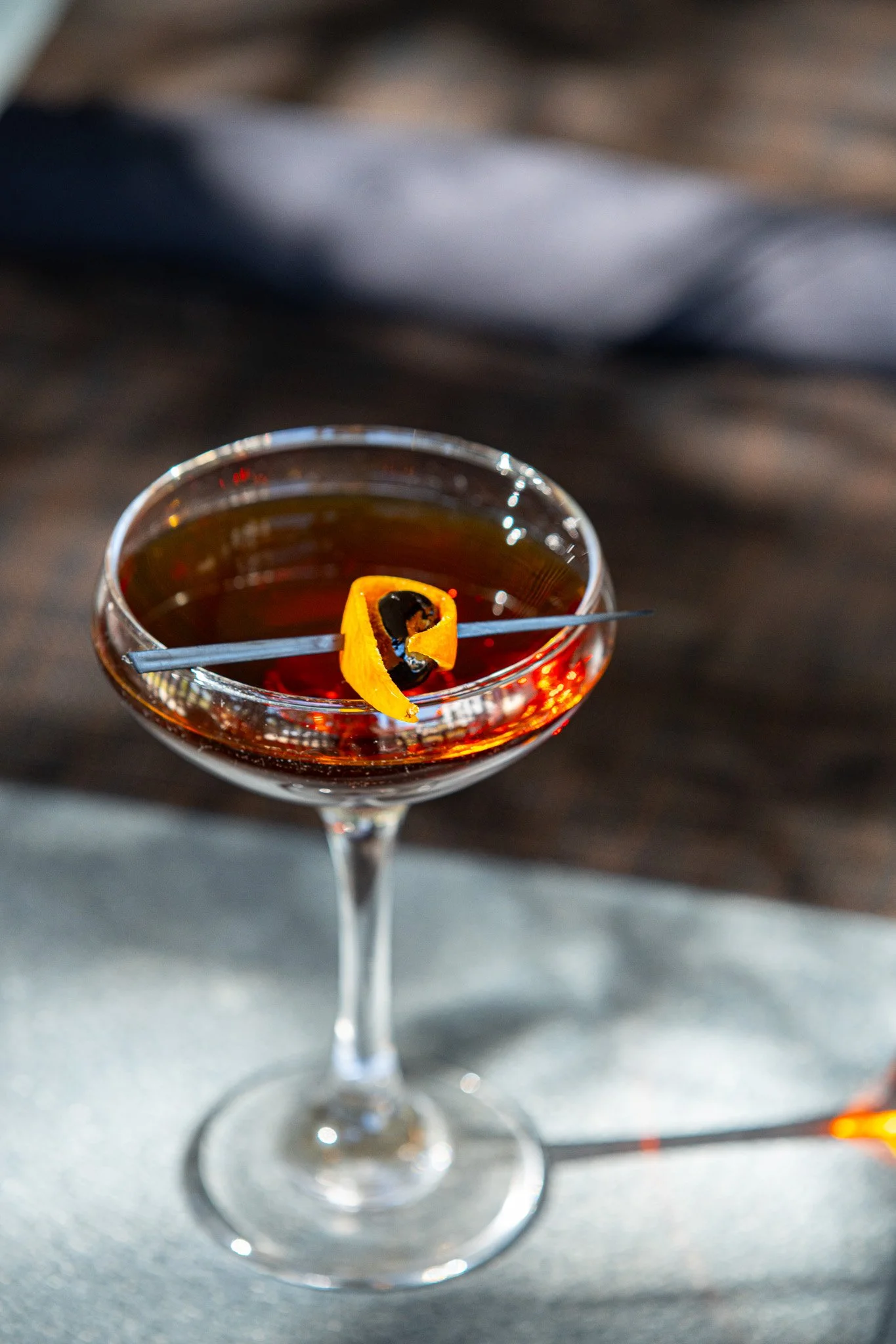A cocktail in a coupe glass garnished with an orange twist and a cherry, on a reflective surface with a blurred dark background.