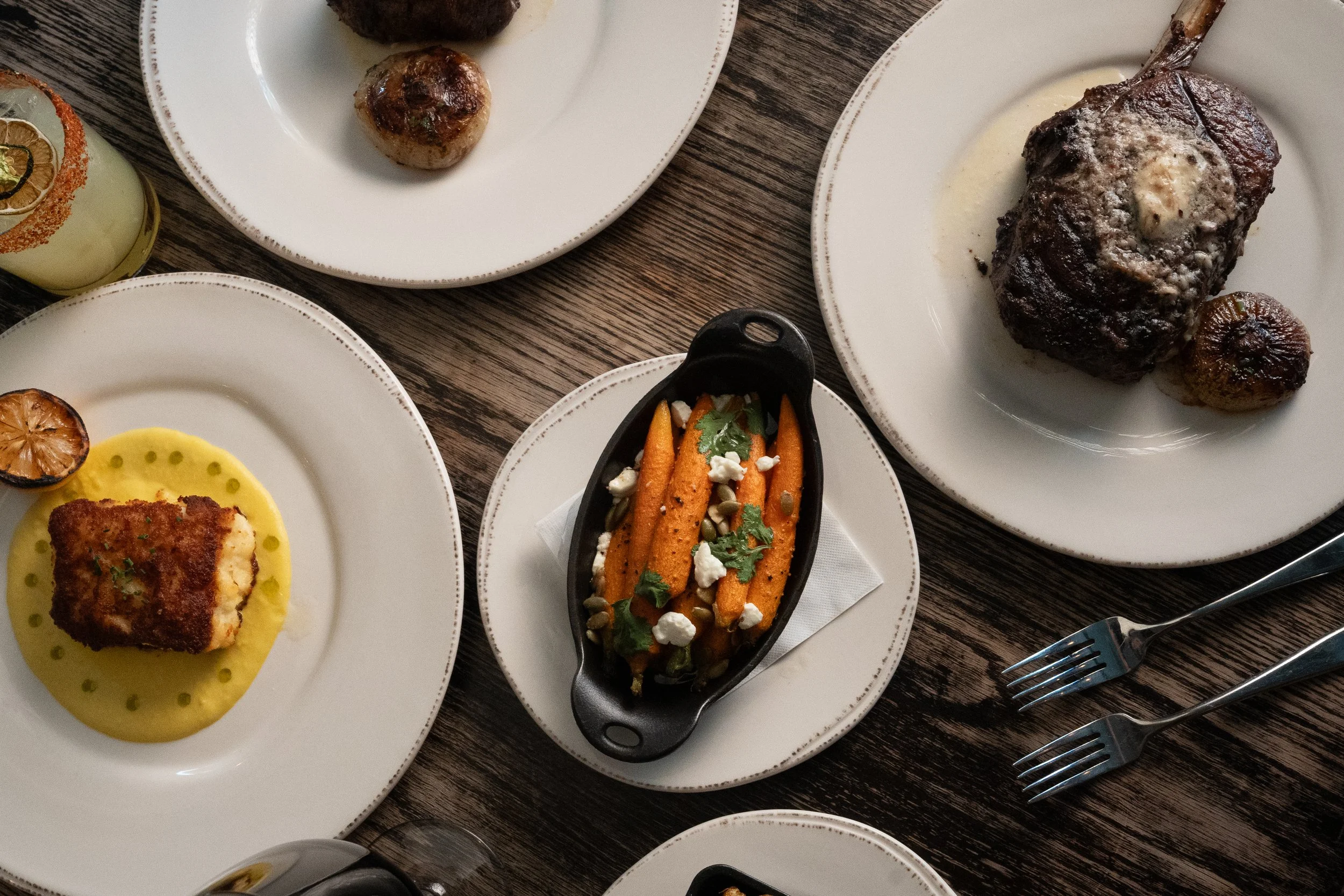 An overhead view of a table with various plated dishes, including roasted meat with vegetables, stuffed fish or chicken, roasted vegetables, and other entrees, set on white plates on a wooden table with metal utensils.