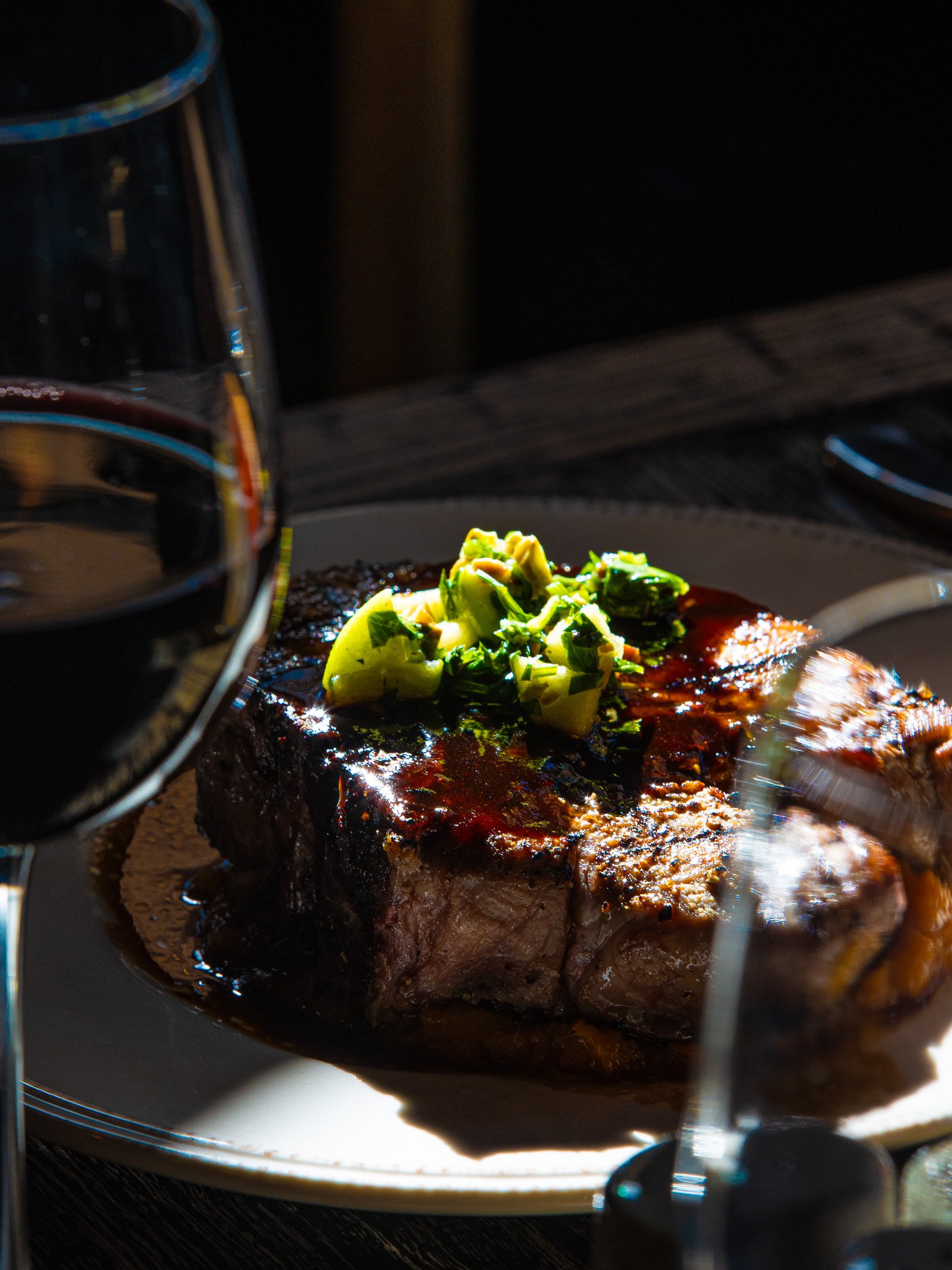 A plated grilled steak topped with chopped green onions, with a glass of red wine nearby.