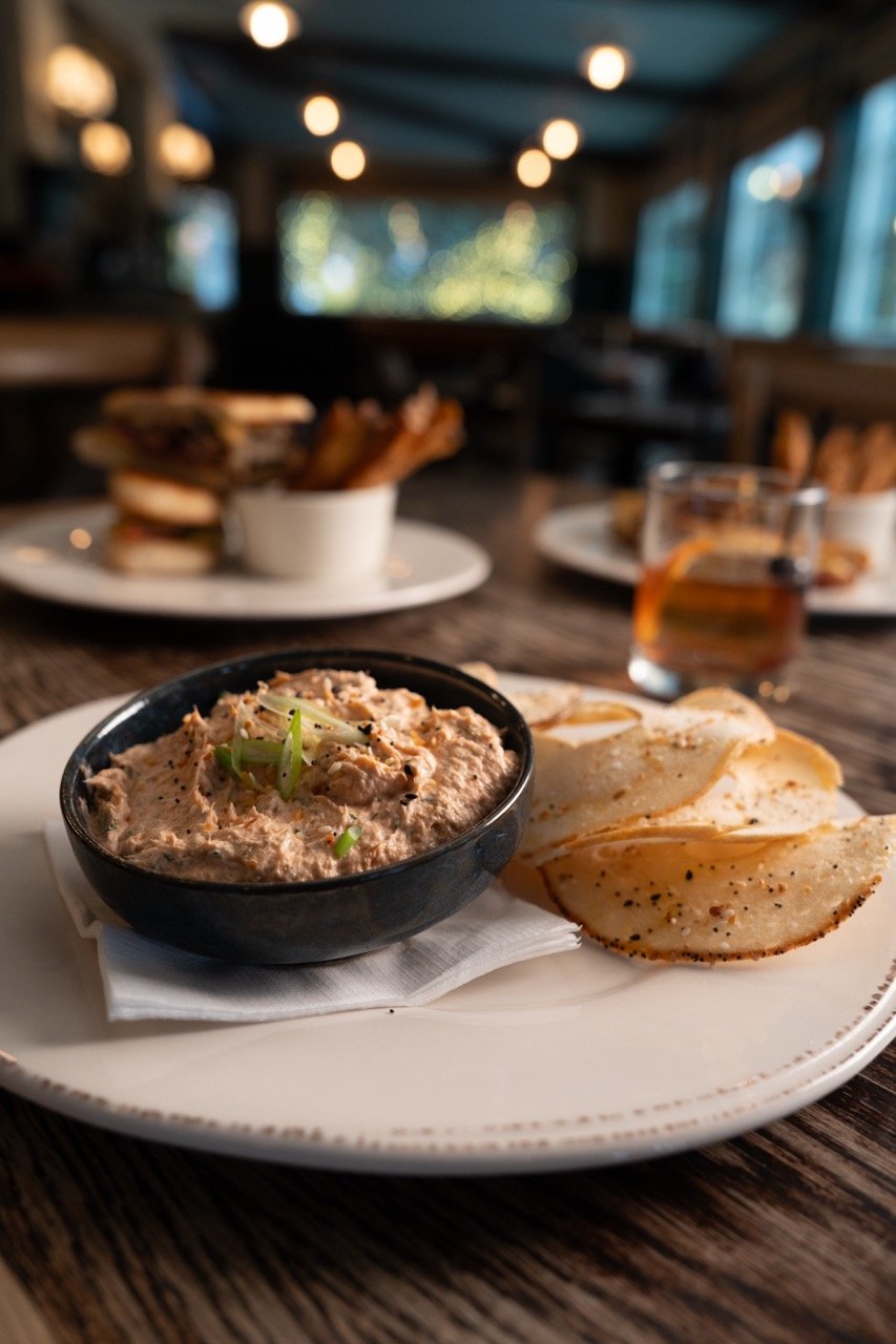 A plate with potato chips, a bowl of creamy tuna salad garnished with green onions, and a glass of liquor, on a wooden table in a restaurant.