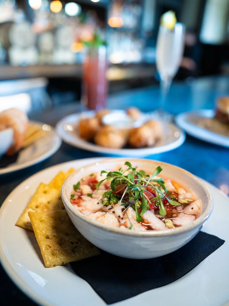 A bowl of shrimp ceviche garnished with microgreens, served with crispy chips on a white plate, with blurred plates and drinks in the background.