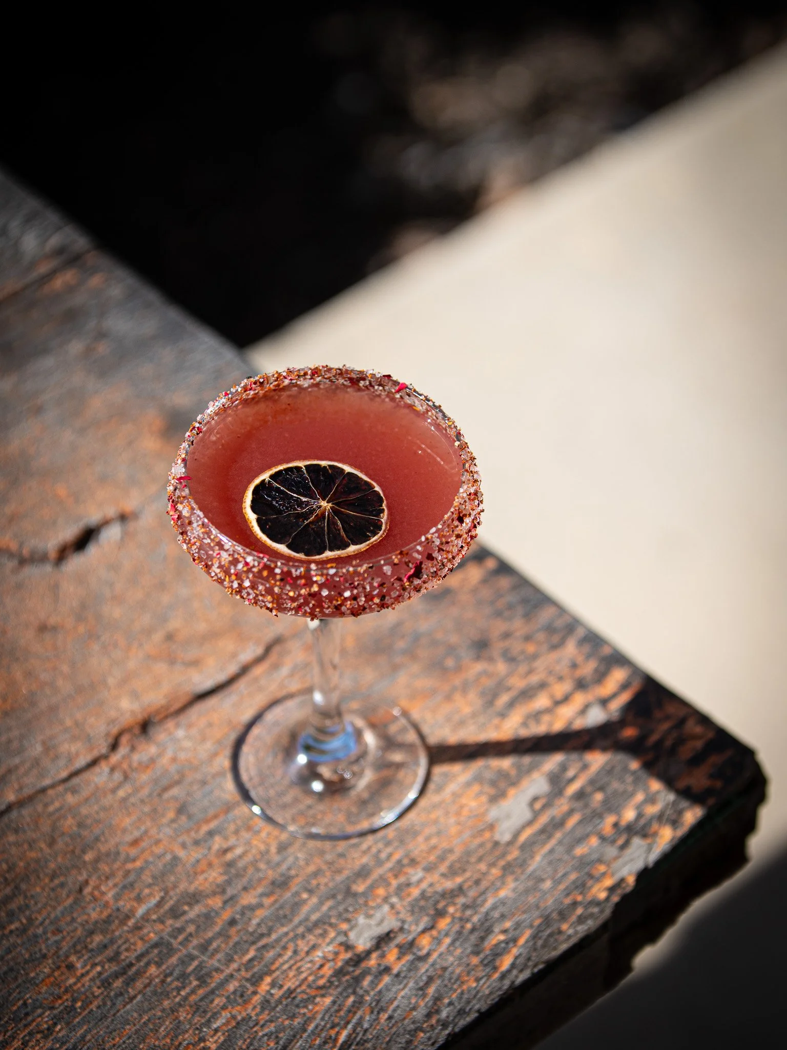 A pink cocktail in a stemmed glass with a salt rim and a dried citrus slice garnish, sitting on a weathered wooden surface.
