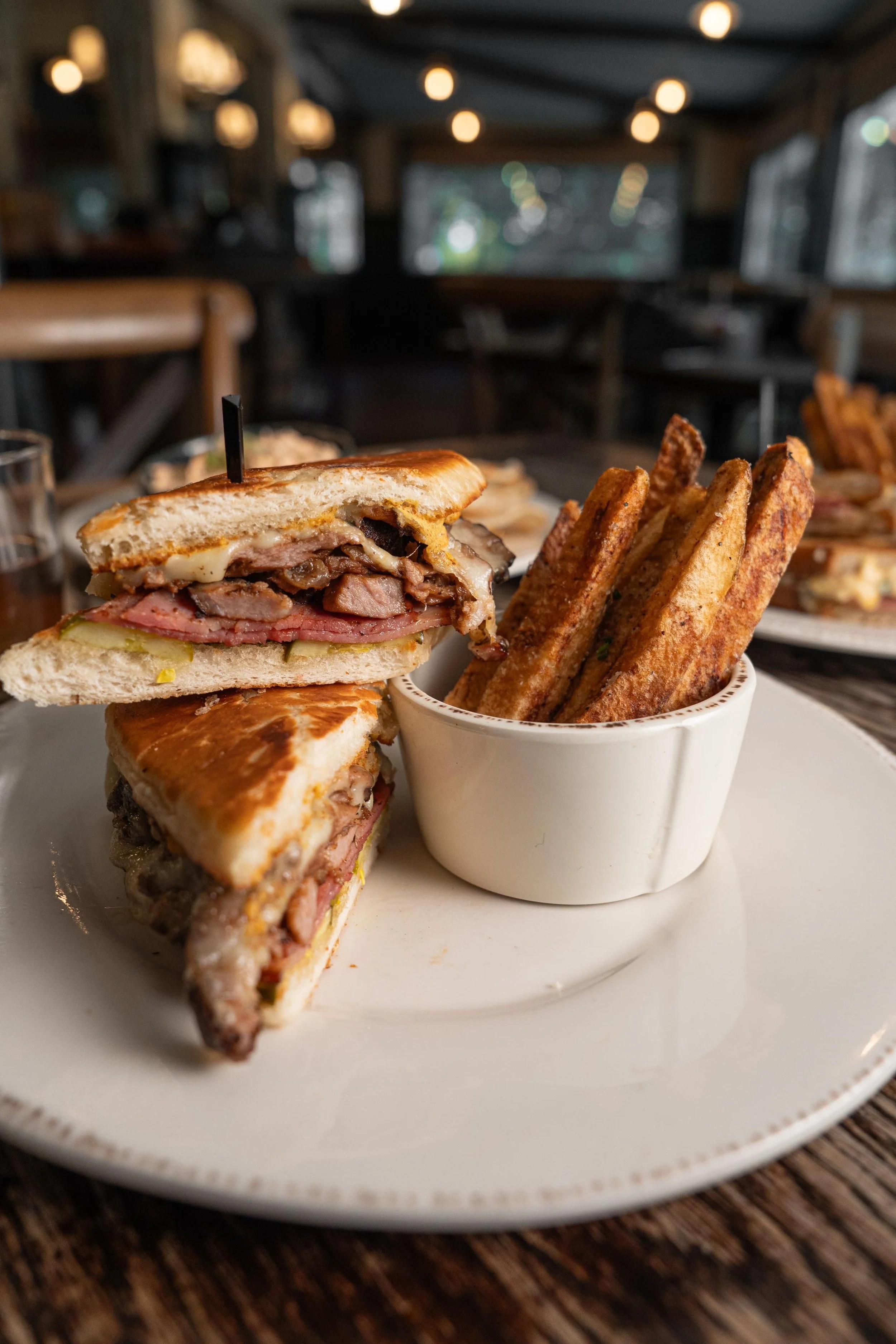A plated sandwich with layers of sliced meats, cheese, and bread, served with fried potato pieces in a white cup