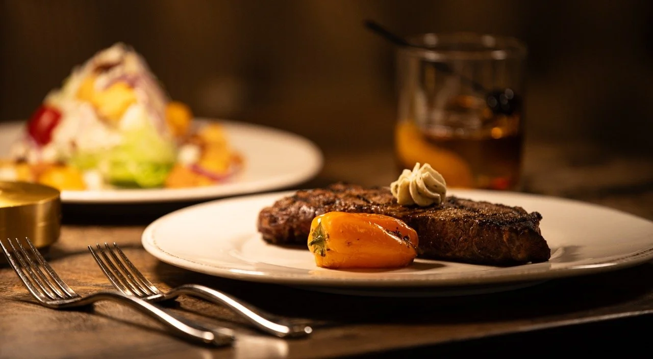 A plate of grilled steak with a dollop of butter and a roasted bell pepper, with a side salad in the background and a glass of dark beverage.