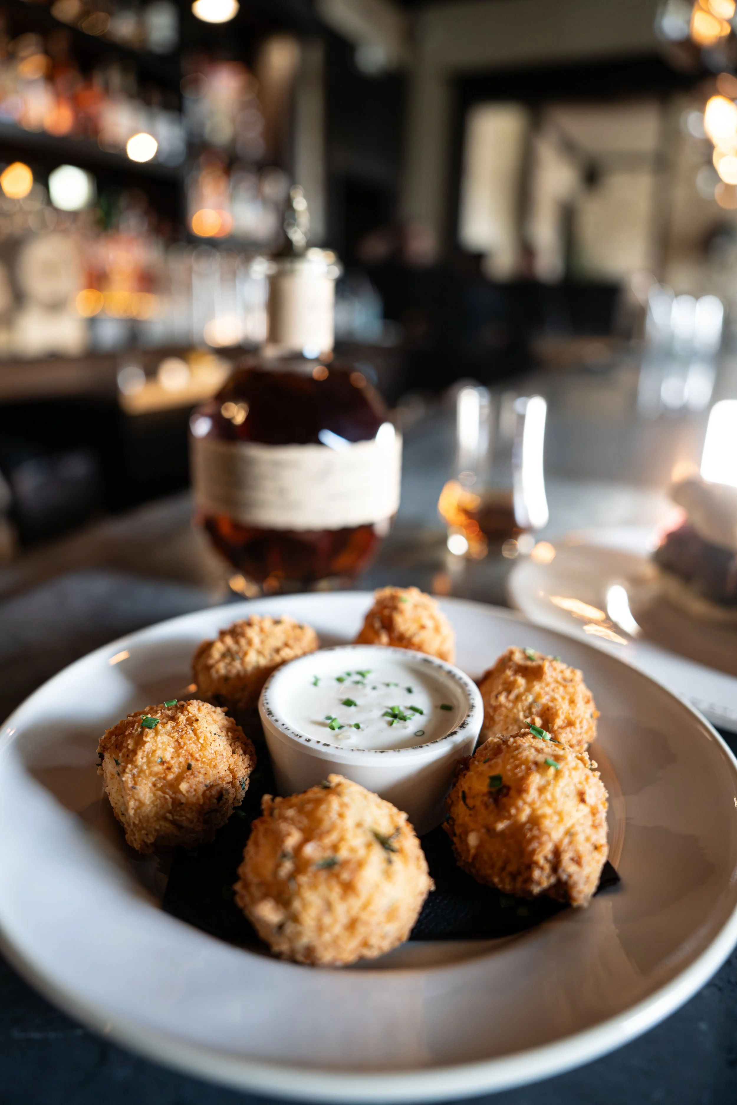 A white plate of fried food balls, garnished with chopped herbs, and a small bowl of white dipping sauce on top. The background appears to be a cozy restaurant with blurred warm lighting and shelves.